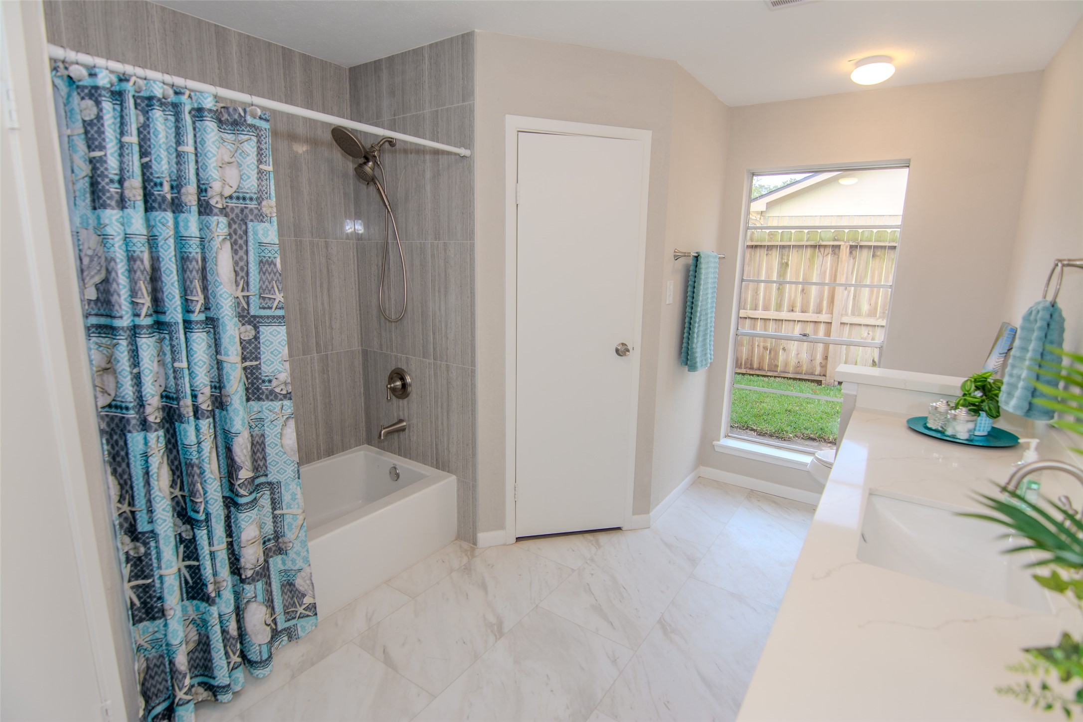 6703 Stonewater Houston, TX 77084 - Photo 19 of 30 Bright and modern bathroom with a large window providing natural light, featuring a tub and shower combination with a stylish blue curtain. The room includes a dual-sink vanity and has a neutral color palette, complemented by white tiles and chrome fixtures.