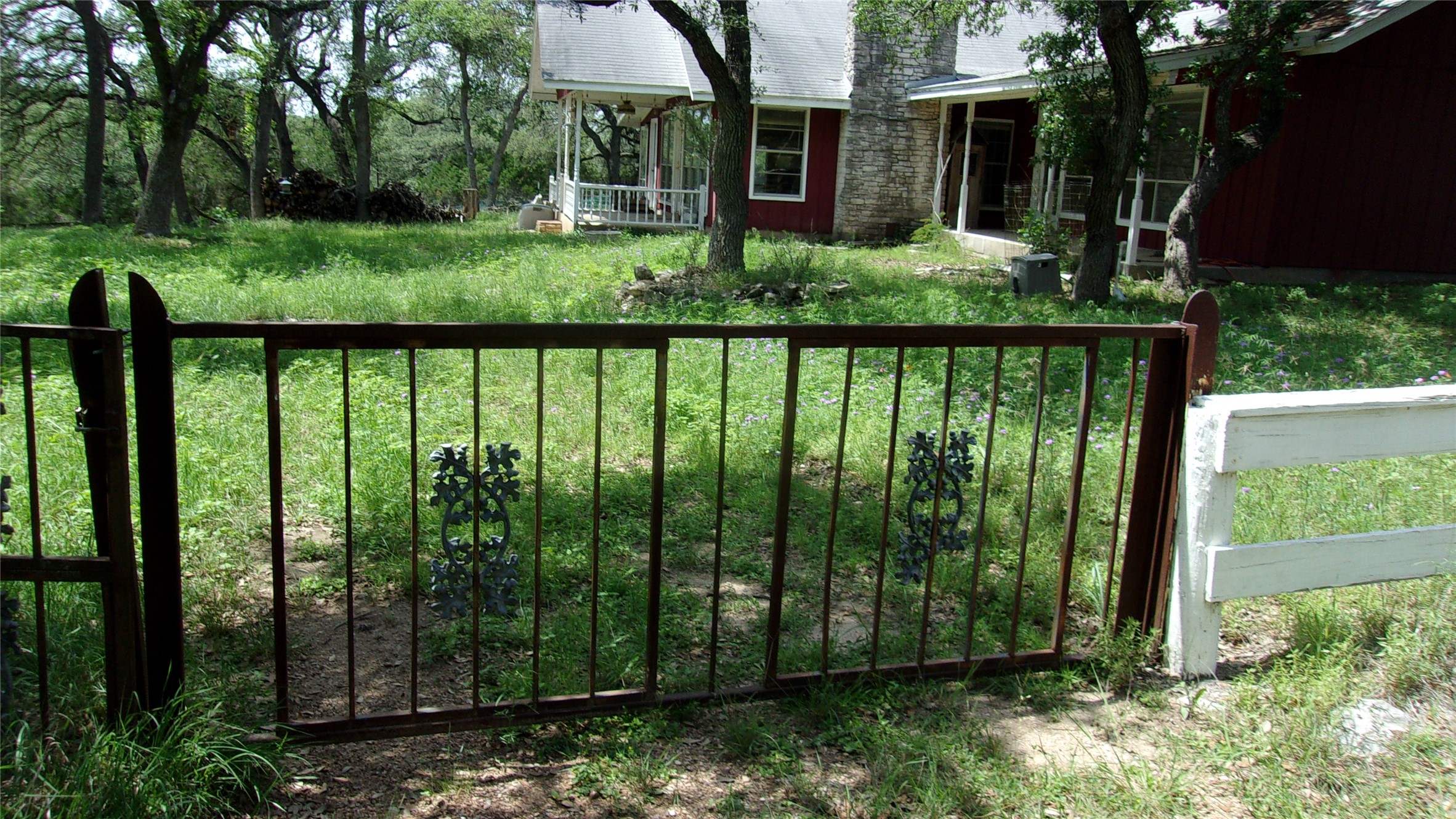 702 Cottonwood Creek Road Dripping Springs, TX 78620 - Photo 3 of 31 Ornate wrought iron gate entry to the property, featuring a white wooden fence post and a stone-clad exterior building with a covered porch in the background