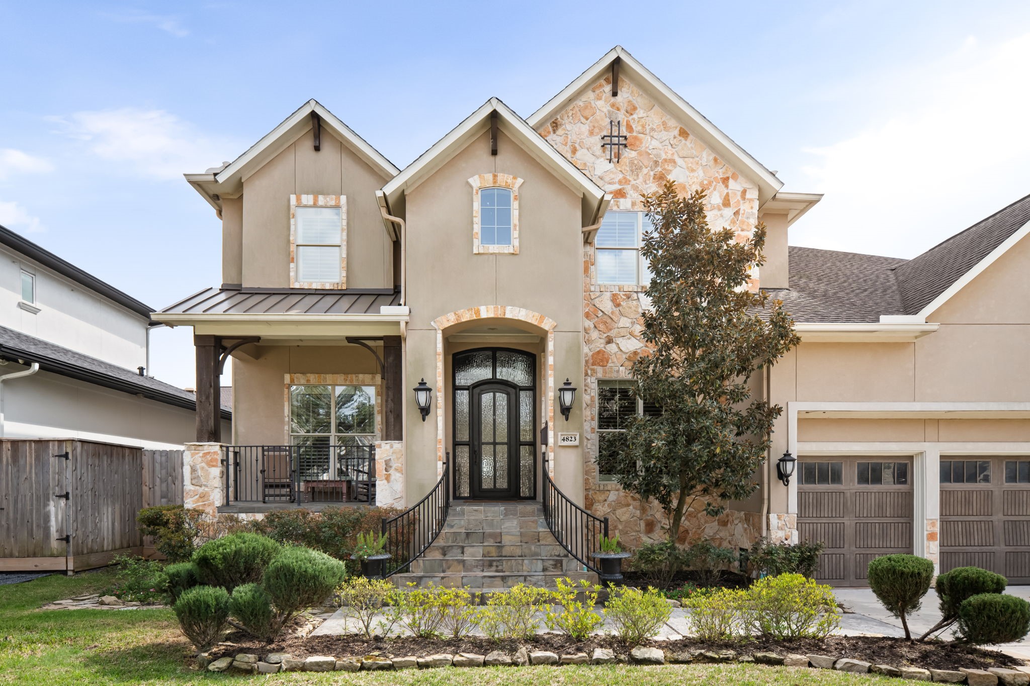 This is a charming two-story home with a stone and stucco exterior, featuring a welcoming front porch and arched entryway. The well-maintained landscaping includes trimmed shrubs and a small tree, complementing the three-car garage.