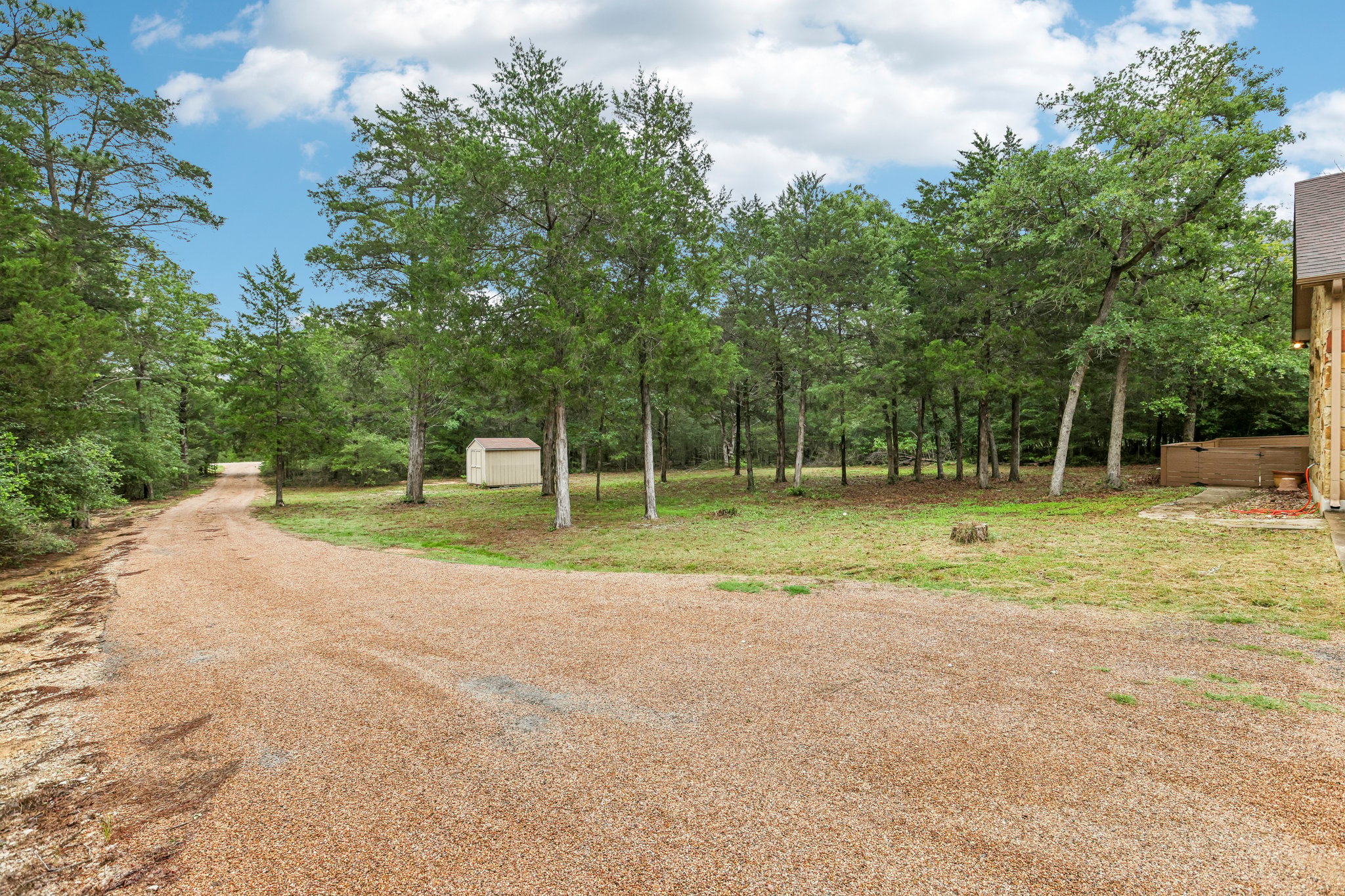 267 Corral Road Smithville, TX 78957 - Photo 4 of 40 Gravel driveway with a tan storage shed, mature trees, and a partial exterior view of the main structure
