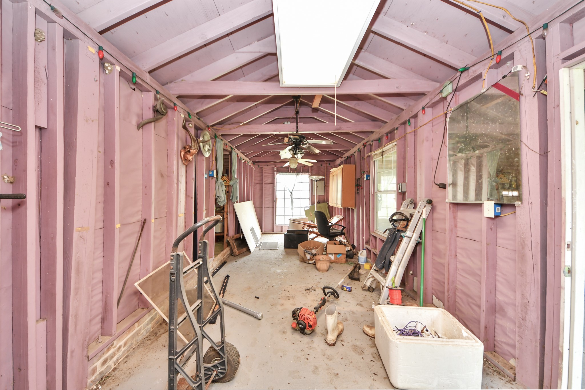 12746 Roy Road Pearland, TX 77581 - Photo 36 of 47 This photo shows a long, narrow utility or storage room with exposed beams and walls painted in a light purple. The space contains various tools and equipment, with a ceiling fan and fluorescent lighting. Natural light enters through a window at the far end.