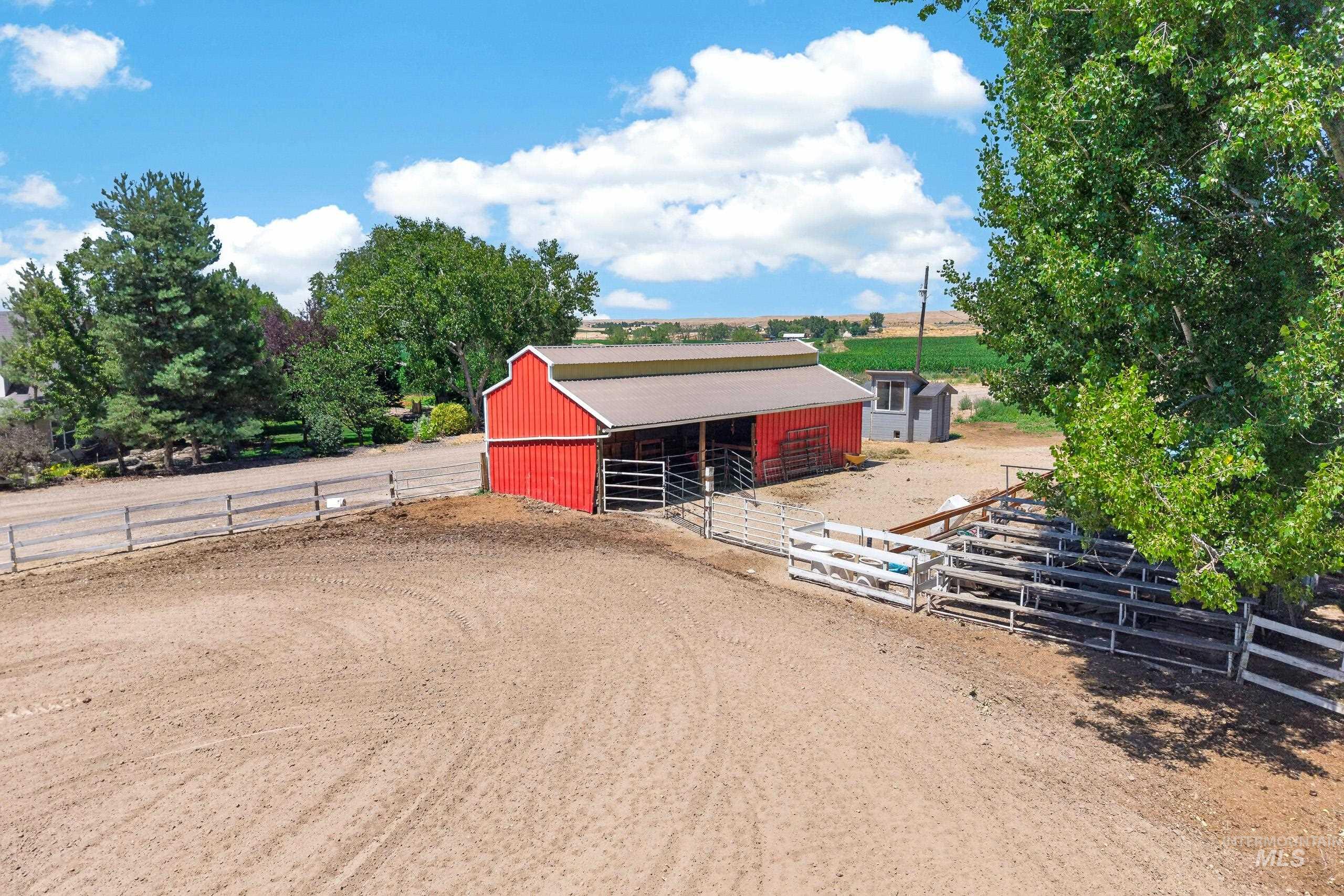 9696 Gloria Road Middleton, ID 83644 - Photo 41 of 50 Red barn structure with a metal roof and open stalls, surrounded by a fenced corral and an expansive dirt arena