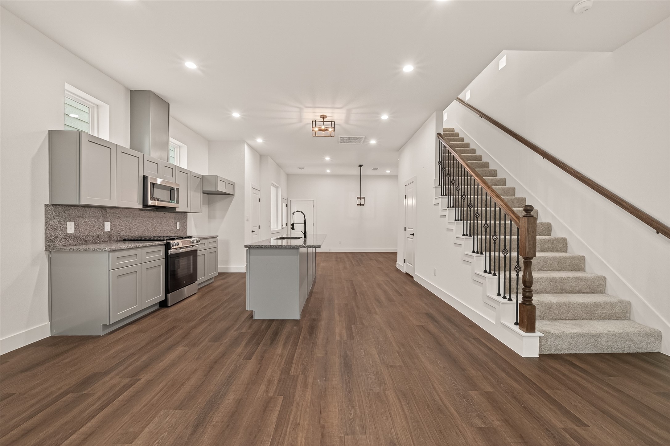 6808 Madrid Street Houston, TX 77021 - Photo 14 of 31 This photo showcases a modern, open-concept kitchen and living area with sleek gray cabinetry, stainless steel appliances, and a large island. The space features warm wood flooring and a carpeted staircase with elegant railing, leading to the upper level. Bright and inviting, it offers a spacious setting for family gatherings and entertaining.