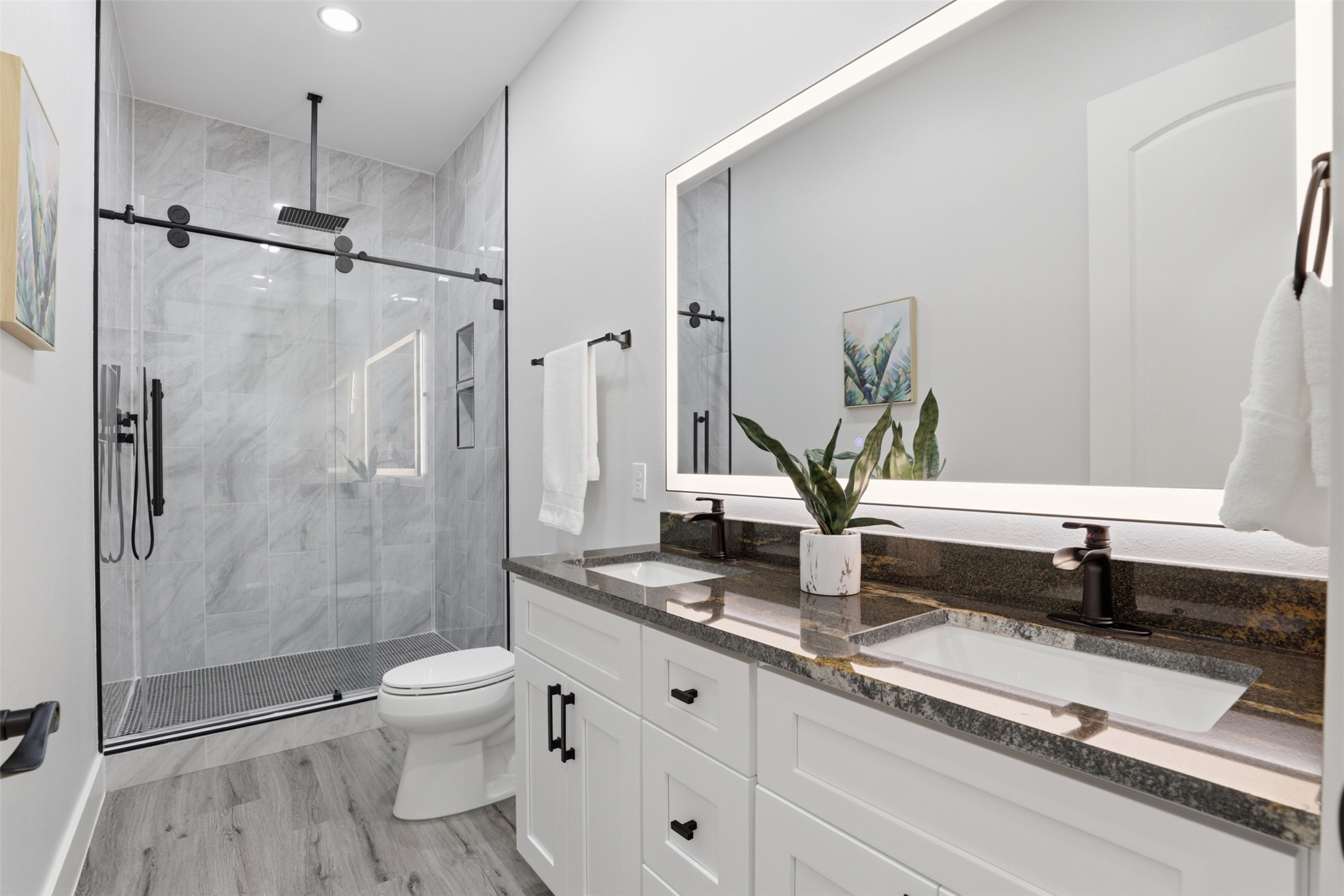 123 Los Robles Road Elgin, TX 78621 - Photo 19 of 21 Contemporary bathroom featuring a dual vanity with dark countertops and undermount sinks, an illuminated mirror, a glass-enclosed shower with a rain shower head and matte black fixtures, and wood-finish flooring