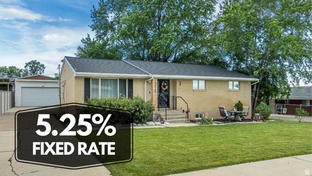 Brick exterior residence featuring a dark shingle roof, well-maintained front lawn, mature trees, and a detached garage with a concrete driveway