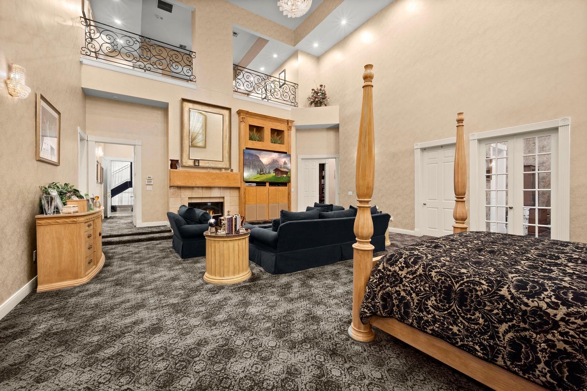 19525 Doerre Road Spring, TX 77379 - Photo 13 of 39 This is a spacious, elegantly appointed primary bedroom with high ceilings, large windows, and an open second-level balcony. It features a comfortable seating area, a fireplace, and patterned carpeting, creating a luxurious and inviting atmosphere.