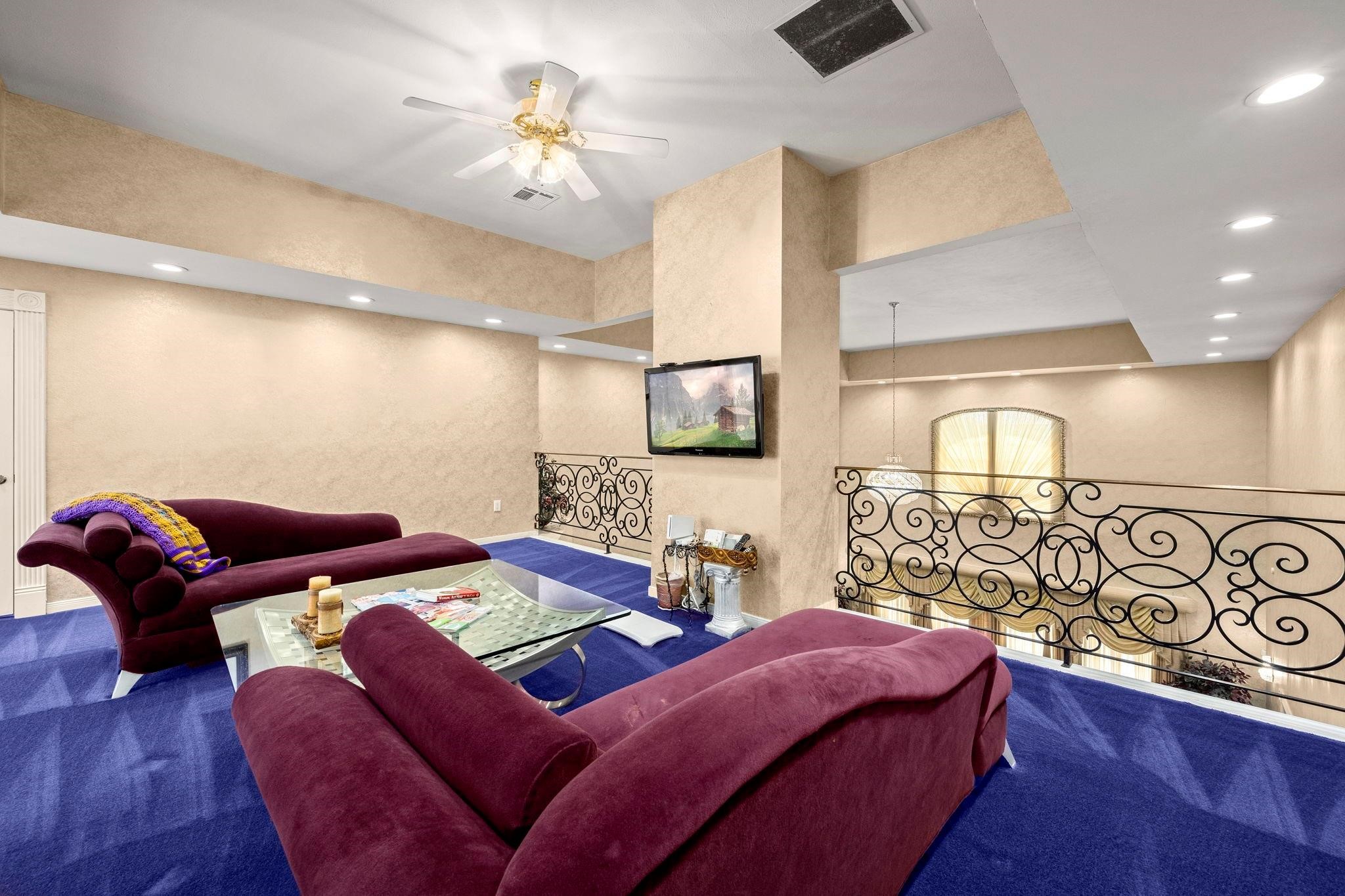 19525 Doerre Road Spring, TX 77379 - Photo 14 of 39 This is a spacious, primary bedroom, balcony area featuring plush maroon sofas, a glass coffee table, blue carpeting, and a decorative wrought iron railing. The room includes recessed lighting, a ceiling fan, and a mounted flat-screen TV.
