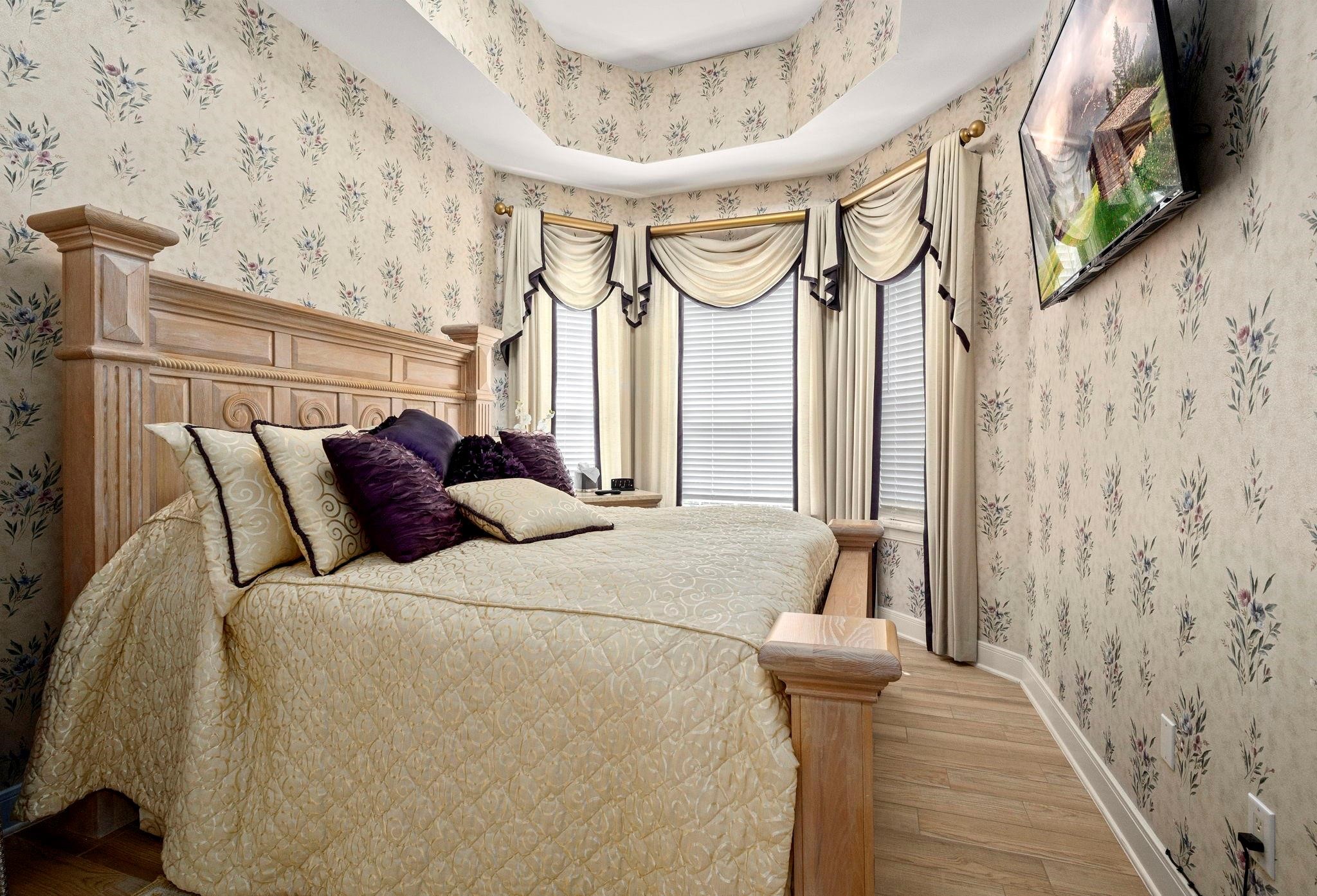 19525 Doerre Road Spring, TX 77379 - Photo 15 of 39 This is a traditional-style bedroom featuring floral wallpaper, a wooden headboard, elegant window draperies, and a matching bedspread with decorative pillows. The room has a classic charm and includes a flat screen T.V. on the wall.