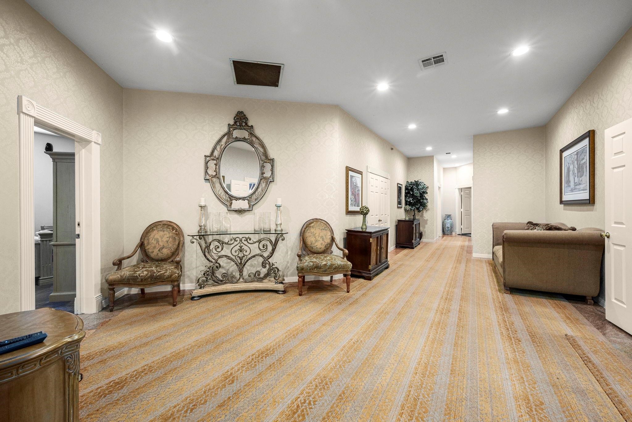 19525 Doerre Road Spring, TX 77379 - Photo 21 of 39 This is a spacious, well-lit hallway with elegant decor, featuring patterned wallpaper, recessed lighting, and plush carpeting. The area is furnished with antique-style chairs, a decorative mirror, and a console table, providing a sophisticated ambiance. Multiple doorways suggest easy access to adjoining rooms.