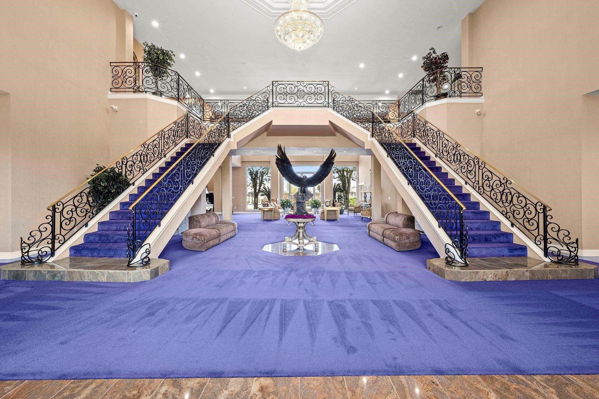 19525 Doerre Road Spring, TX 77379 - Photo 5 of 39 This is a grand foyer with dual sweeping staircases, ornate wrought-iron railings, and a bold purple carpet, leading to an elegant second-floor landing. The space is open and airy, featuring a high ceiling with a chandelier, enhancing the luxurious feel of the home.