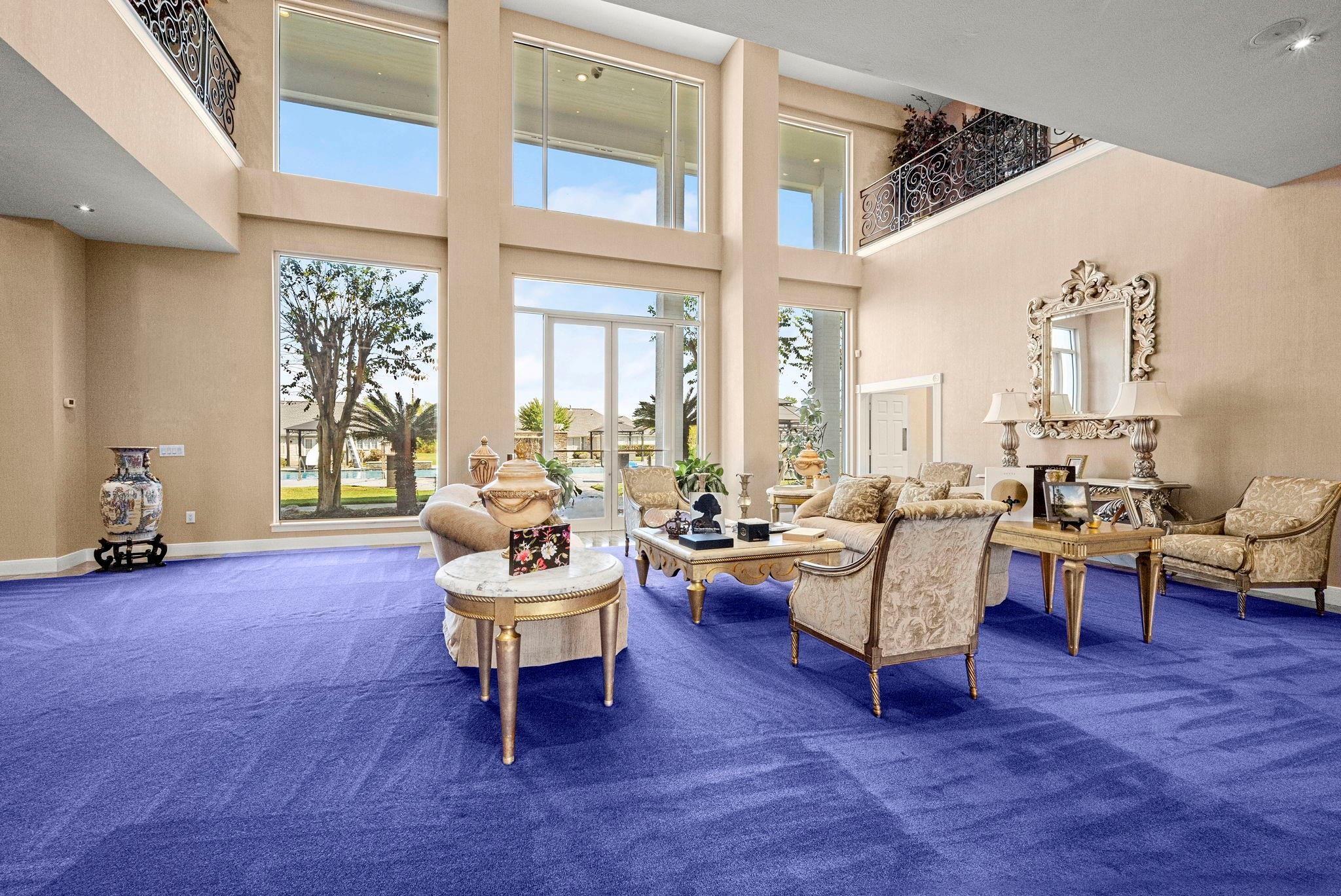 19525 Doerre Road Spring, TX 77379 - Photo 8 of 39 This is a spacious living room with high ceilings, large windows providing ample natural light, and a view of the outdoors. It features a bold purple carpet and classic-style furniture, with a second-story indoor balcony overlooking the space.