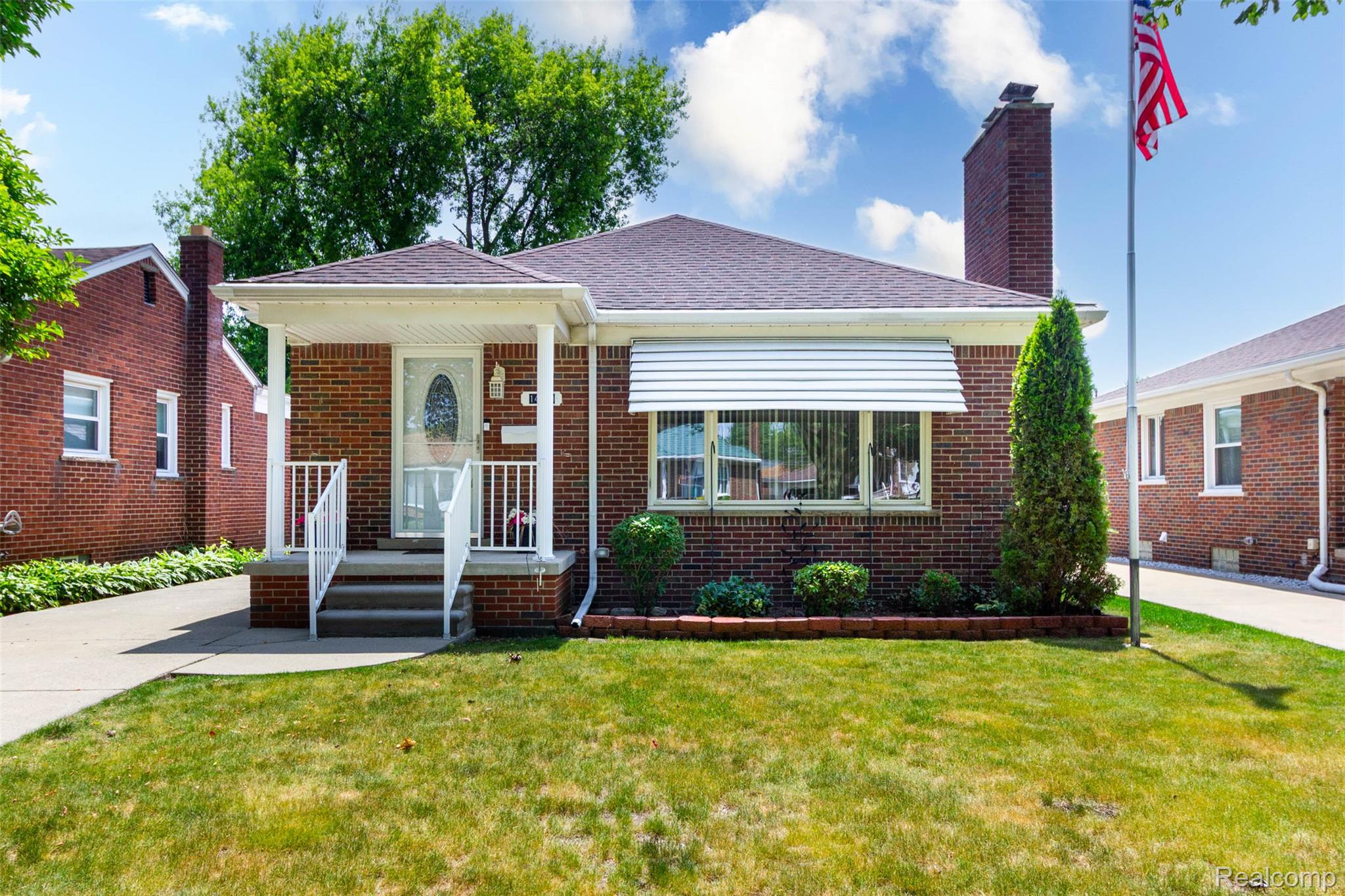Brick exterior featuring a covered porch with white railing, a prominent chimney, and a landscaped front garden bed