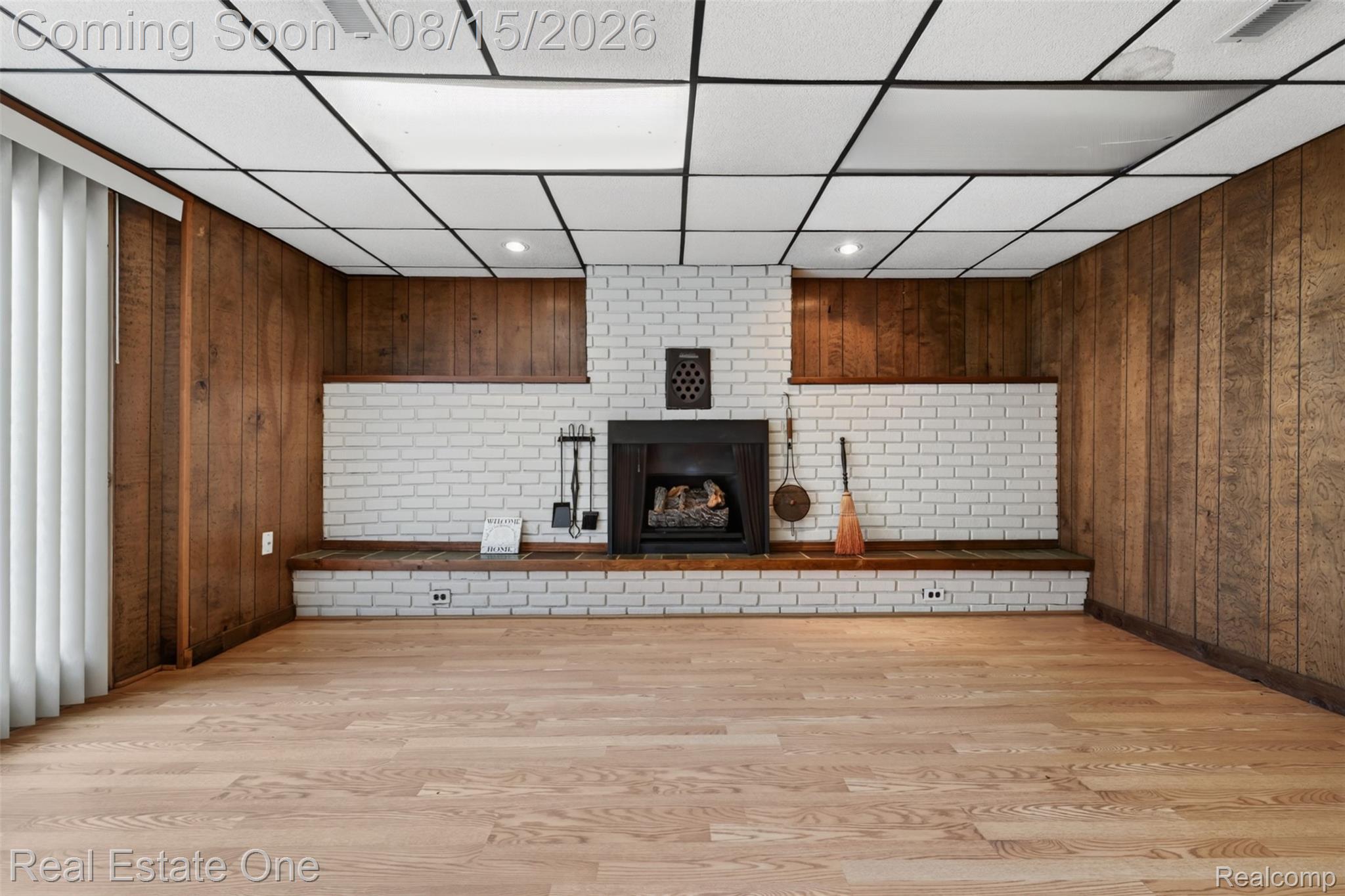 454 South Baldwin Road Oxford, MI 48371 - Photo 35 of 64 Wood-finish flooring, white brick fireplace with black insert, wood paneling, drop ceiling with recessed lighting, and vertical blinds