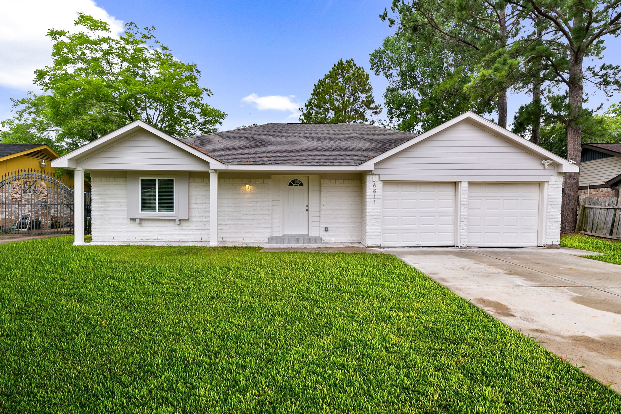 This is a charming single-story home with a clean white exterior, featuring a two-car garage. It is surrounded by a well-maintained lawn and framed by mature trees, providing a welcoming and serene atmosphere.