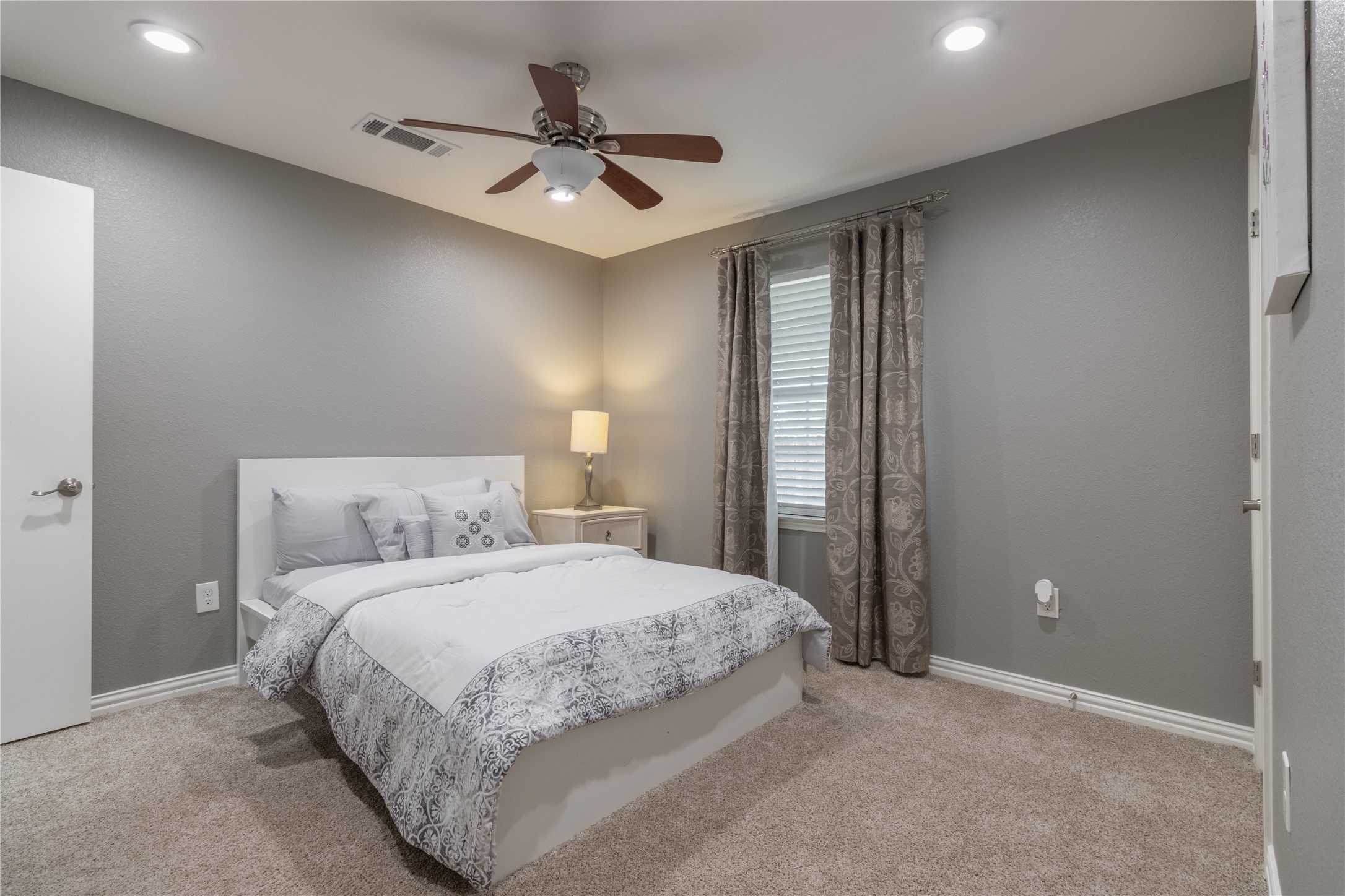17123 Atlanta Street Humble, TX 77396 - Photo 13 of 29 This cozy bedroom features soft gray walls, a plush carpet, and a ceiling fan. It includes a comfortable bed with decorative bedding and a nightstand with a lamp by a window with elegant curtains.