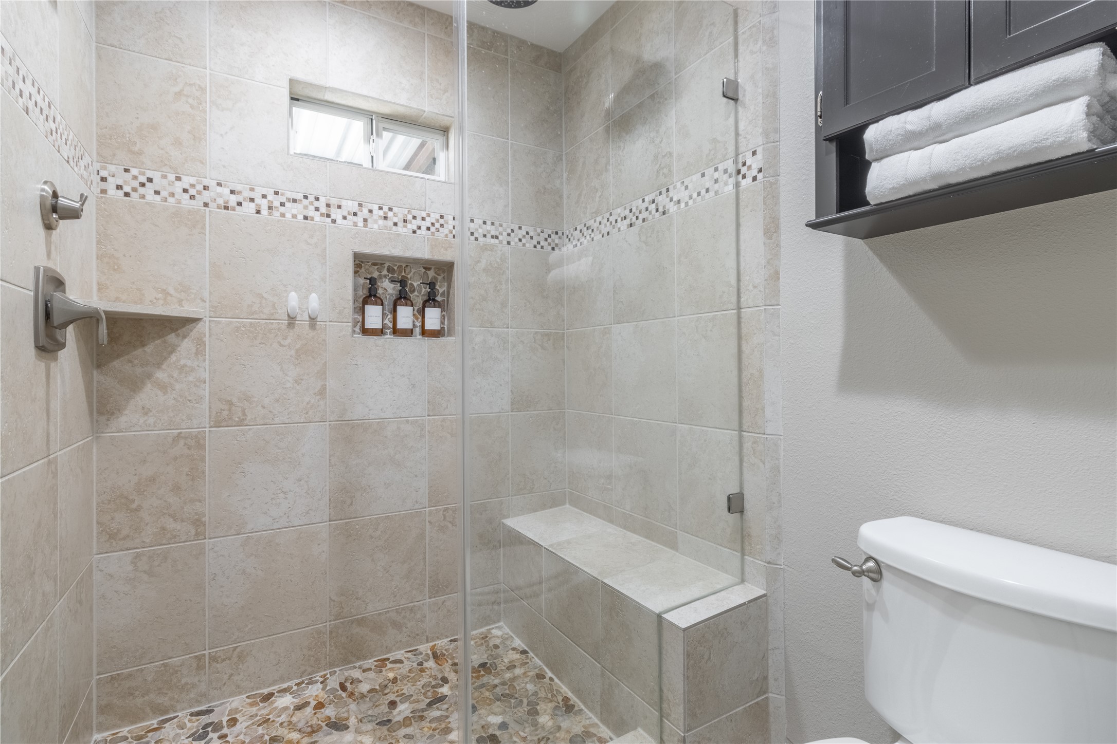 17123 Atlanta Street Humble, TX 77396 - Photo 16 of 29 This bathroom features a modern walk-in shower with elegant beige tiles and a mosaic accent strip. It includes a built-in bench, niche for toiletries, and pebble stone flooring. Adjacent to the shower is a neatly organized storage shelf with towels.