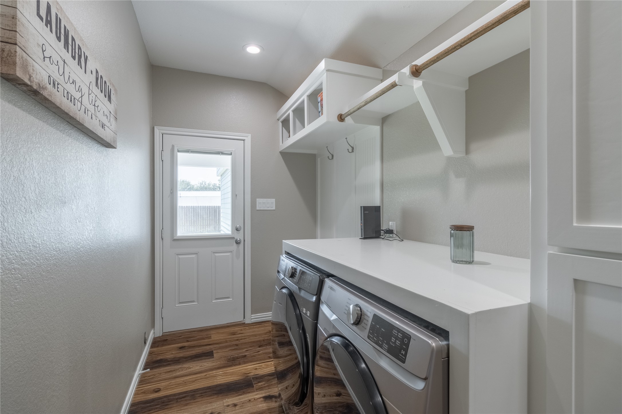 17123 Atlanta Street Humble, TX 77396 - Photo 23 of 29 This laundry room features modern appliances, a wooden floor, and ample storage with overhead shelving and hooks. It includes a door with a window for natural light and a decorative "Laundry Room" sign.
