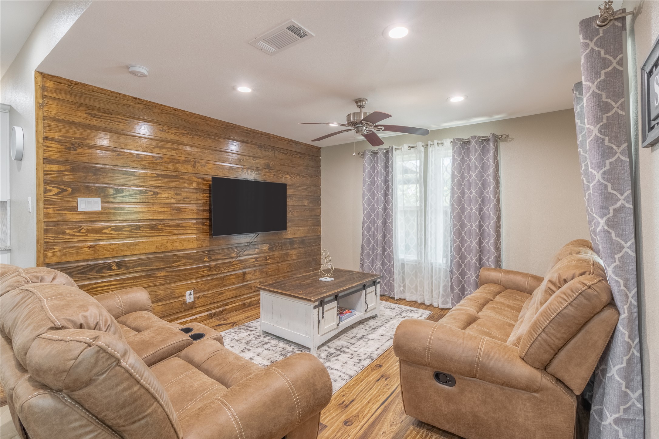 17123 Atlanta Street Humble, TX 77396 - Photo 4 of 29 This cozy living room features a rustic wood accent wall with a mounted TV, plush reclining sofas, a stylish ceiling fan, and large windows with patterned curtains for natural light.