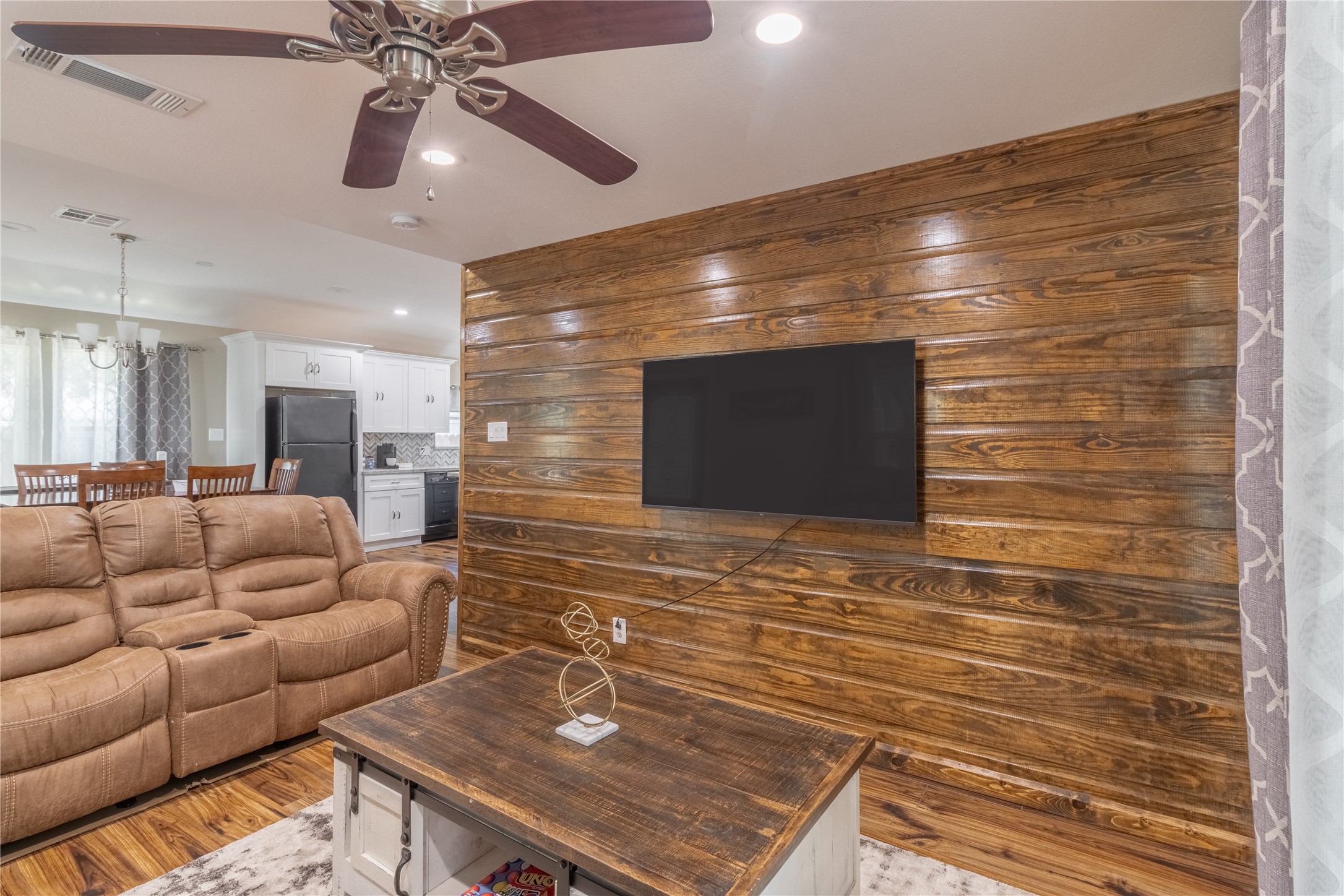 17123 Atlanta Street Humble, TX 77396 - Photo 5 of 29 Cozy living area with a rustic wooden accent wall featuring a mounted TV. The space includes a comfortable recliner sofa, a ceiling fan, and a view into a bright kitchen with modern appliances and dining area. Perfect for relaxed living and entertaining.
