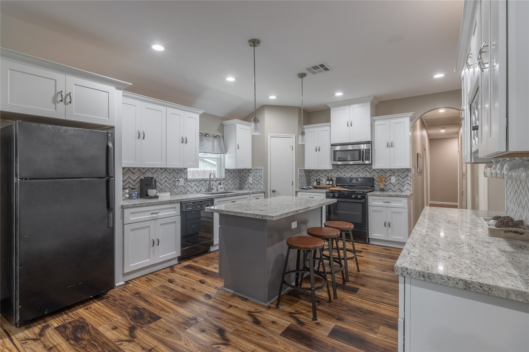 17123 Atlanta Street Humble, TX 77396 - Photo 8 of 29 This kitchen features a modern design with white cabinetry, granite countertops, and a central island with seating. It includes stainless steel appliances, a stylish herringbone backsplash, and warm wood flooring. Ideal for cooking and entertaining.
