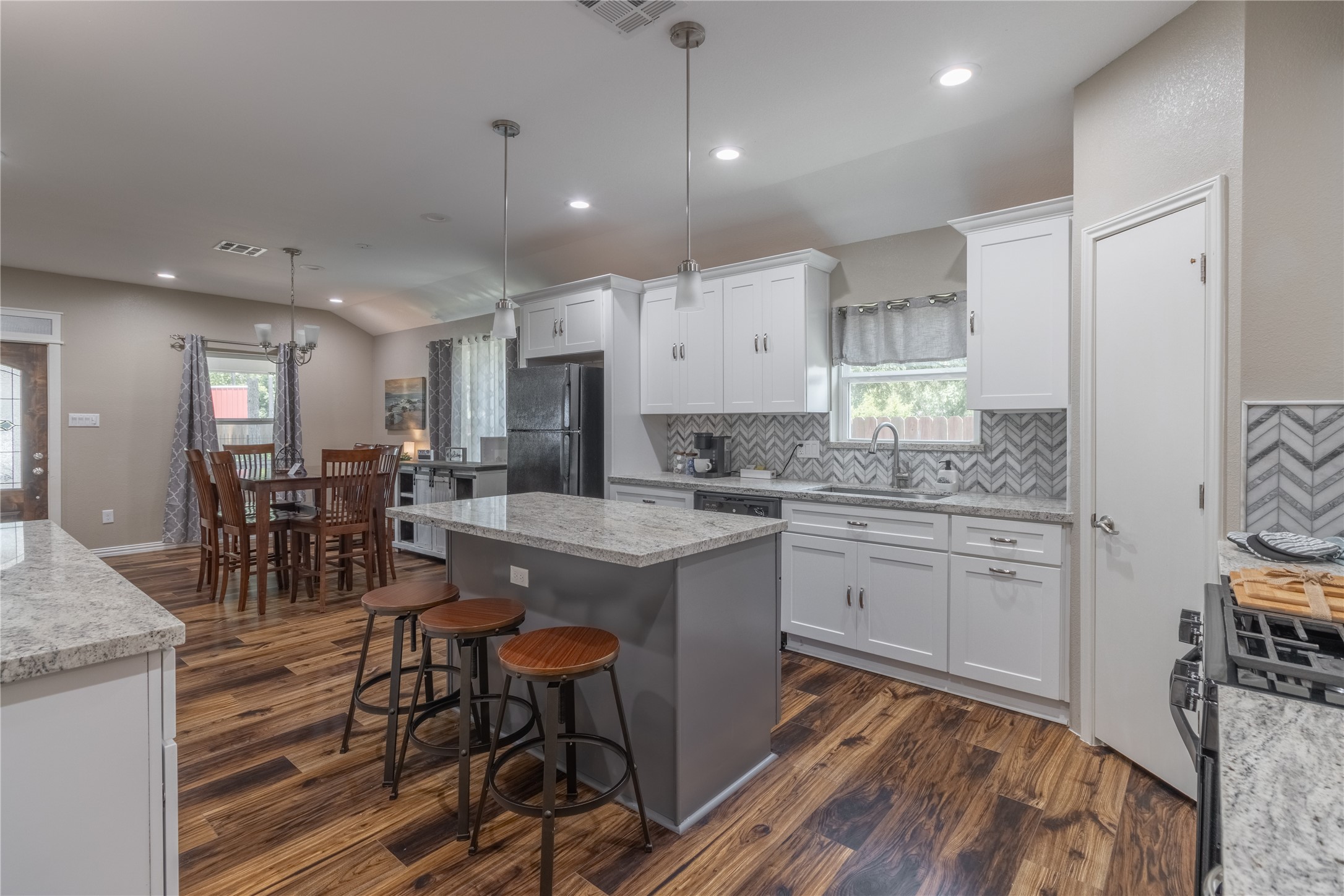 17123 Atlanta Street Humble, TX 77396 - Photo 9 of 29 This modern kitchen features white cabinetry, a large island with seating, and granite countertops. The space includes stainless steel appliances, a stylish herringbone backsplash, and an open layout that flows into a dining area with wood flooring and ample natural light.