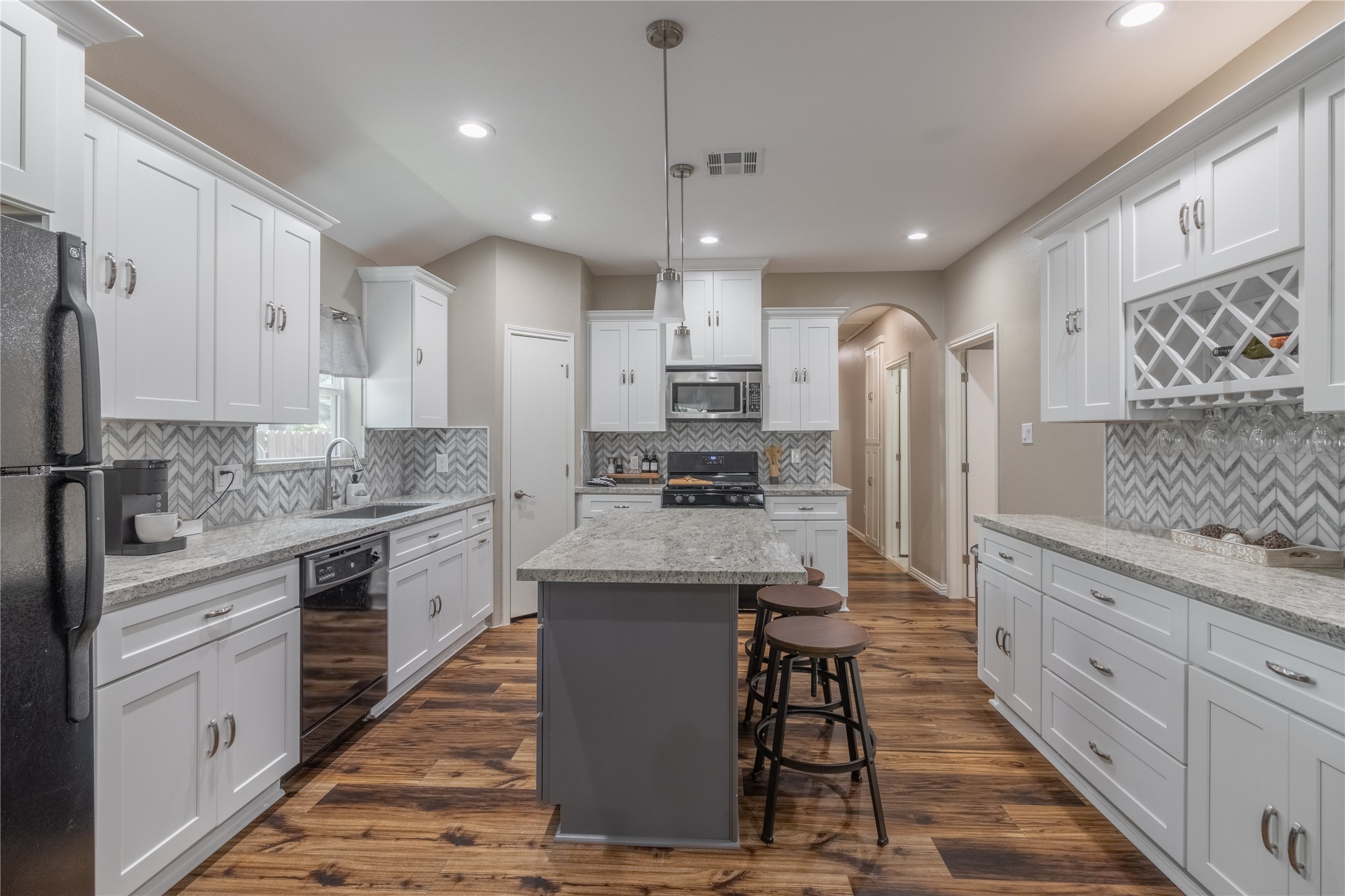 17123 Atlanta Street Humble, TX 77396 - Photo 10 of 29 This kitchen features a modern design with white cabinetry, granite countertops, and a central island with seating. The chevron backsplash adds a stylish touch, while stainless steel appliances offer functionality. Warm wood flooring completes the inviting space.