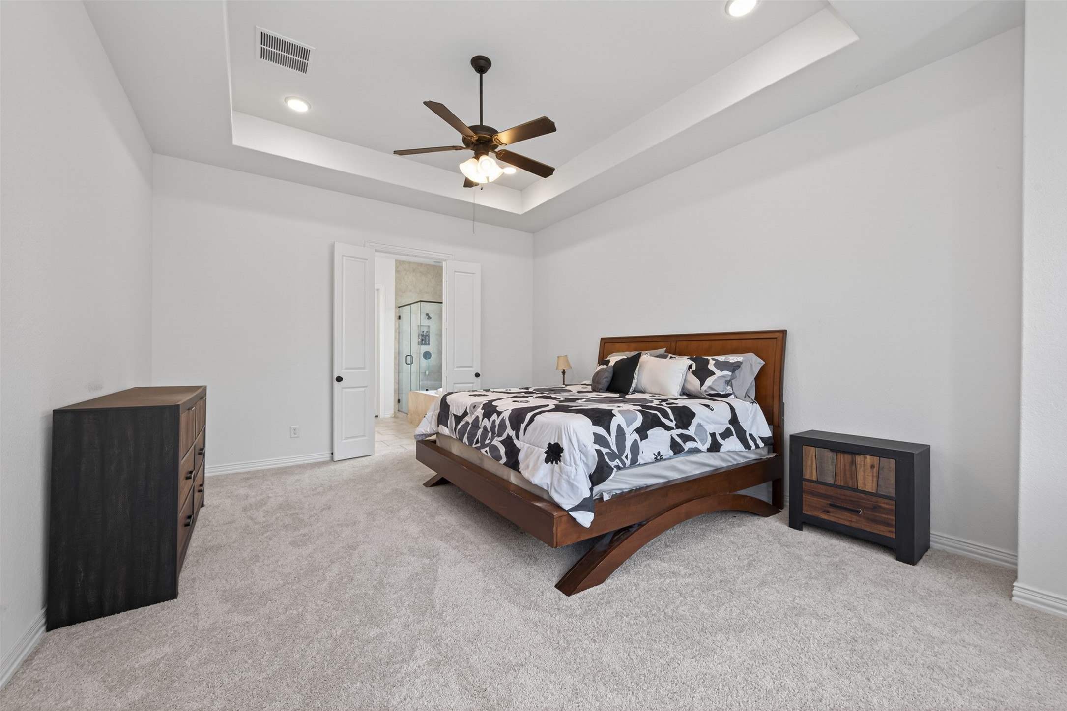 20802 Mirror Point Drive Cypress, TX 77433 - Photo 29 of 37 This exquisite bedroom showcases a contemporary design, complete with tray ceilings and plush carpeting. The space is enhanced by a ceiling fan, recessed lighting, and an adjoining bathroom accessed via double doors. A soothing neutral color palette establishes a tranquil and welcoming ambiance, ideal for relaxation and repose.