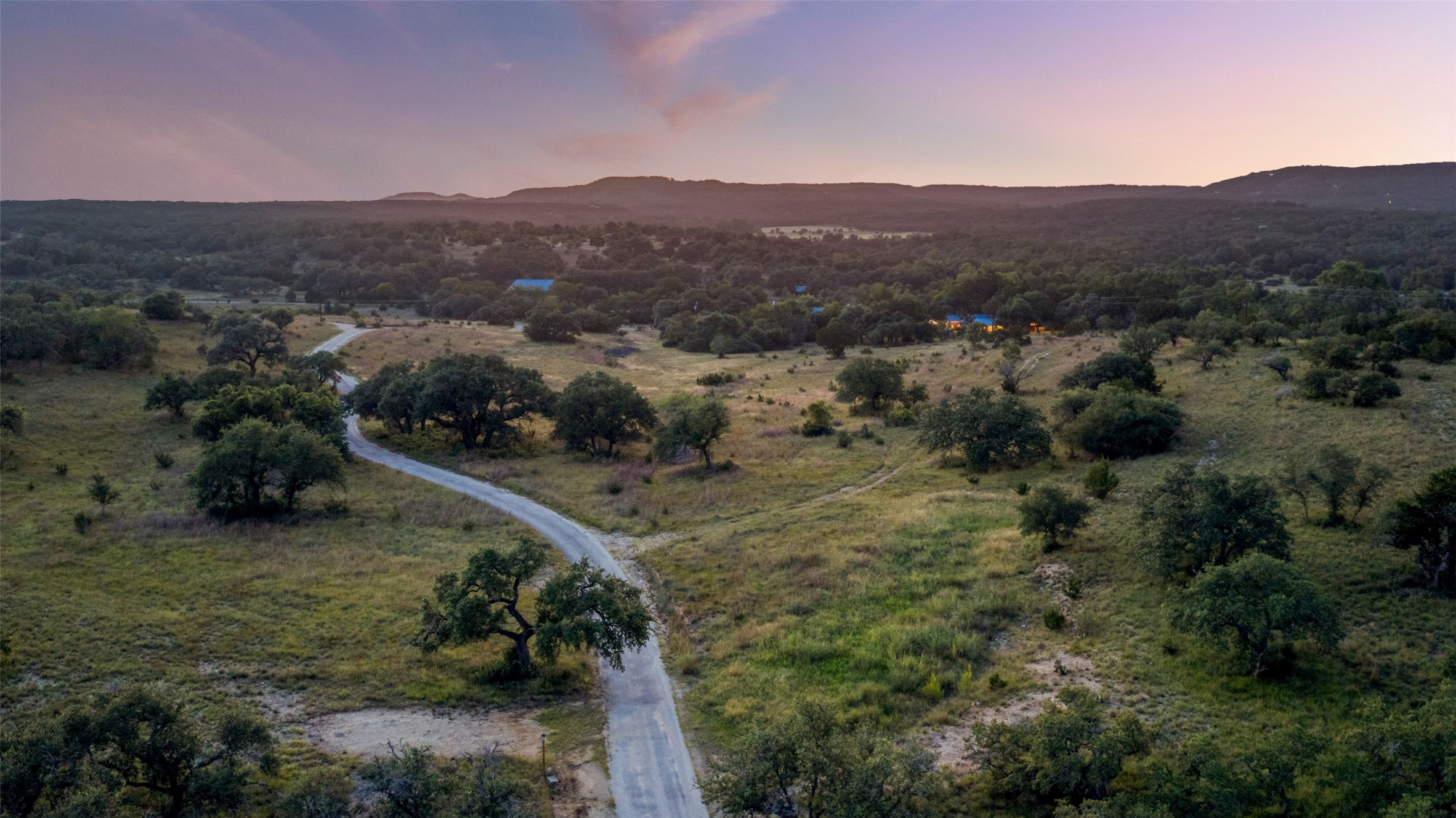 505 Red Corral Ranch Road Wimberley, TX 78676 - Photo 1 of 40 Vast, verdant landscape with a winding driveway leading to distant homes, all under a colorful twilight sky.