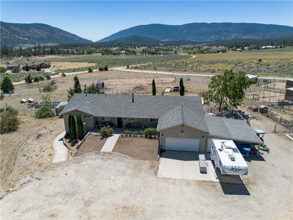 An elevated aerial view highlighting the front exterior of the home, featuring a spacious driveway with ample parking space, an attached garage, landscaped entry paths, and expansive views of the open property stretching toward the mountains.