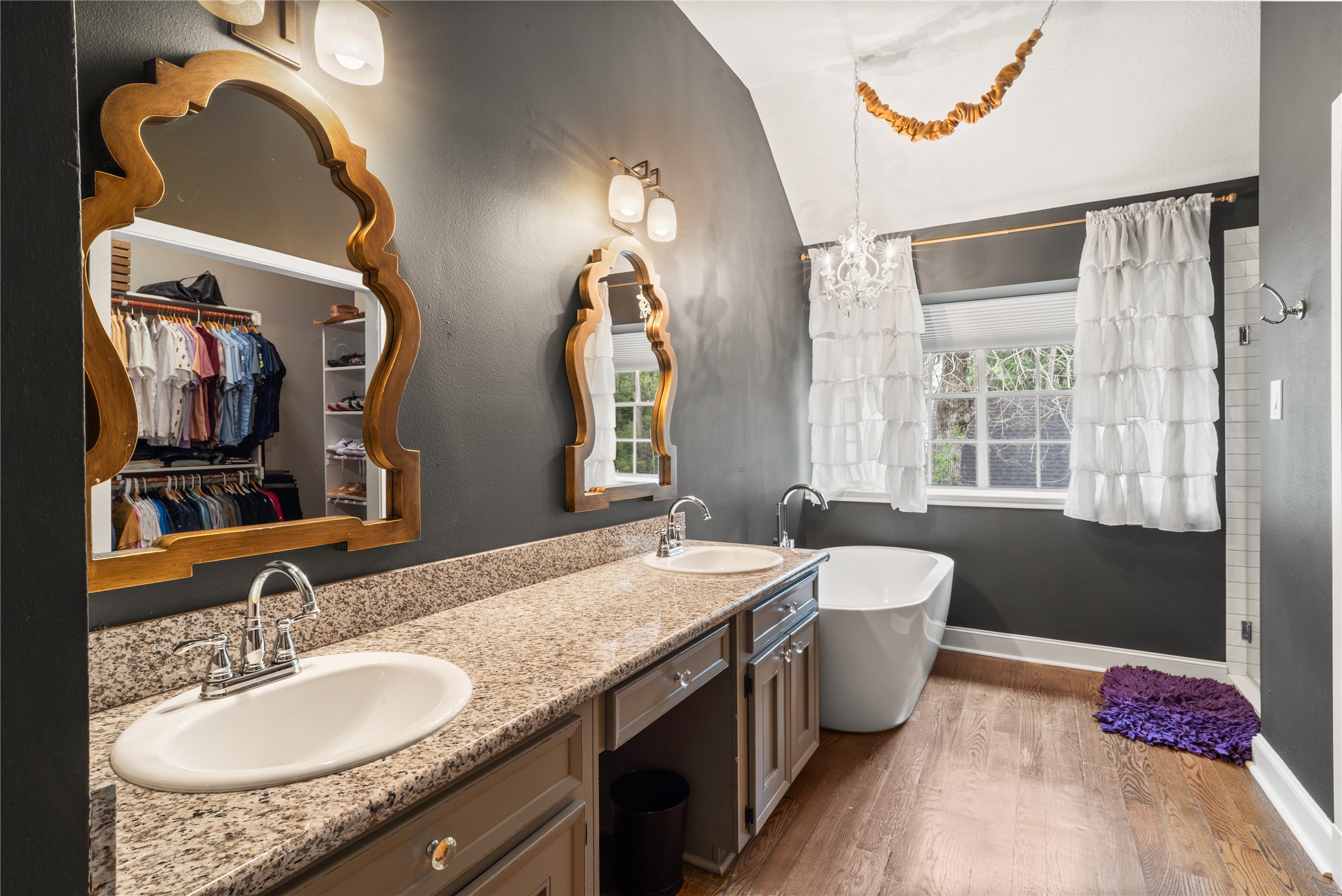 36 Wind River Drive Conroe, TX 77384 - Photo 33 of 45 This stylish bathroom features dual sinks with elegant mirrors, a freestanding bathtub, and hardwood floors. It connects to a spacious walk-in closet, all enhanced by soft lighting and a large window with ruffled curtains.