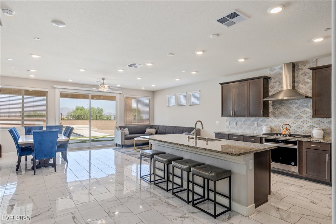5592 Hickory Tree Street Las Vegas, NV 89149 - Photo 27 of 87 Kitchen featuring light marble finish flooring, light stone counters, decorative backsplash, dark brown cabinets, and an island with sink