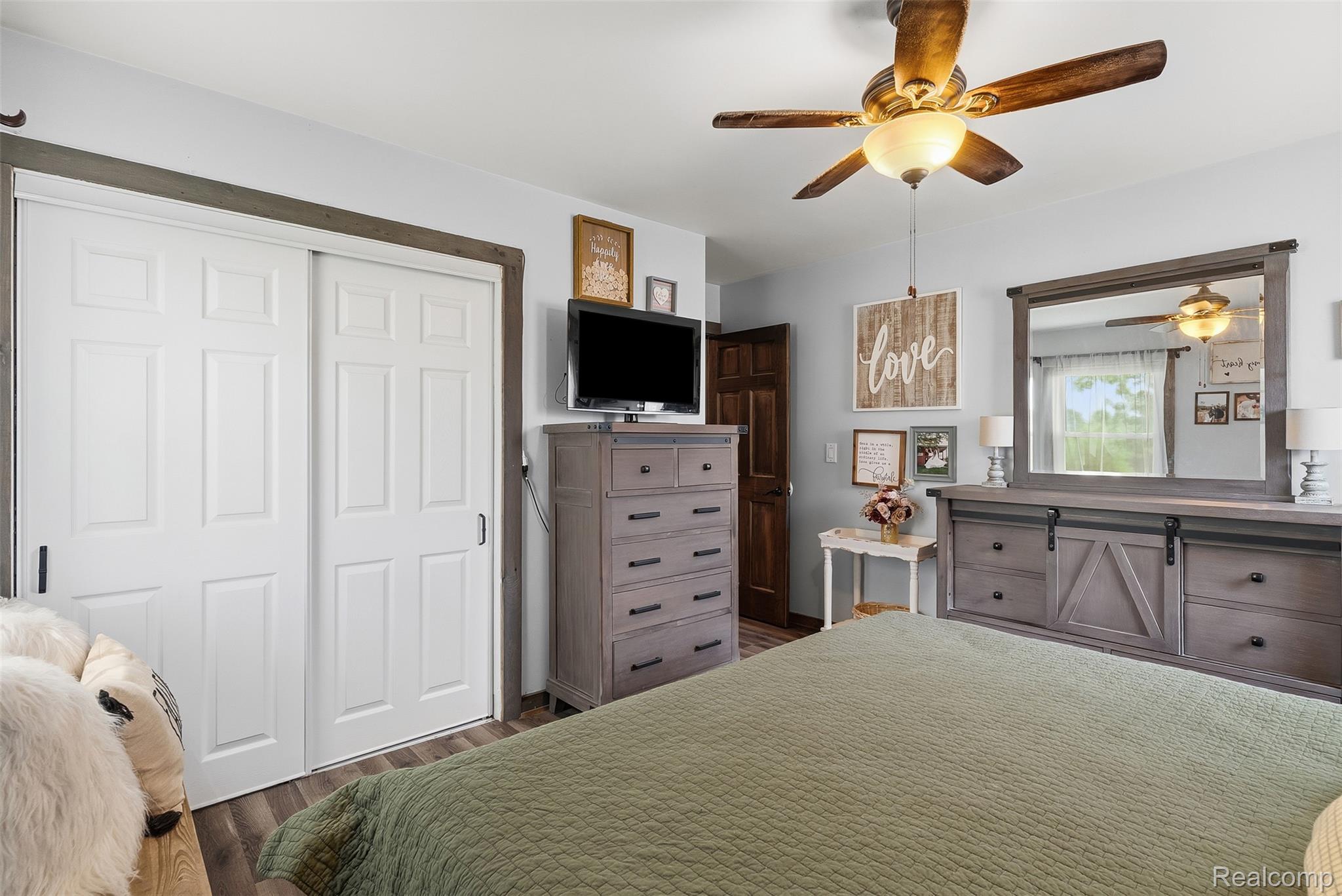 8403 Miller Road Swartz Creek, MI 48473 - Photo 13 of 37 Spacious bedroom featuring light gray walls, wood-finish flooring, a ceiling fan with integrated lighting, a dark wood-paneled door, and white bi-fold closet doors