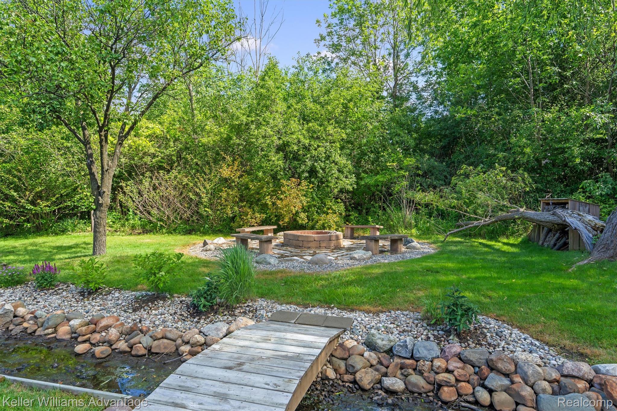 22878 Tree Top Court South Lyon, MI 48178 - Photo 56 of 75 Expansive backyard featuring a brick fire pit, surrounding stone benches, and a decorative wooden footbridge spanning a rock-lined stream bed