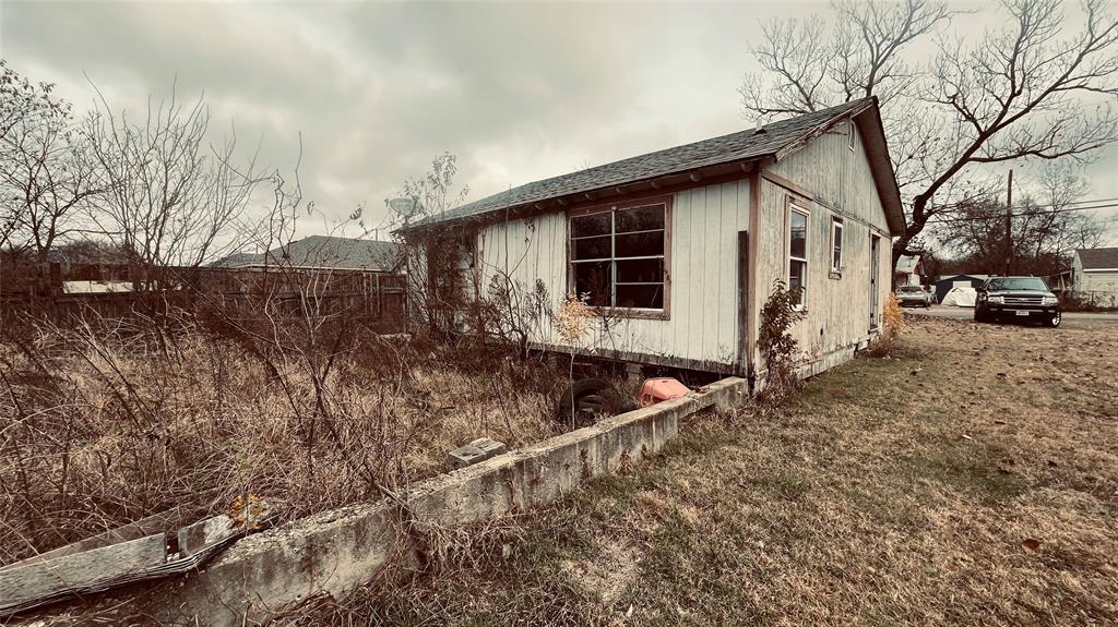 1445 Elk Creek Road Dallas, TX 75253 - Photo 4 of 4 Side and rear view of deteriorated home with broken windows, missing siding, and overgrowth. Yard shows signs of neglect and disrepair.