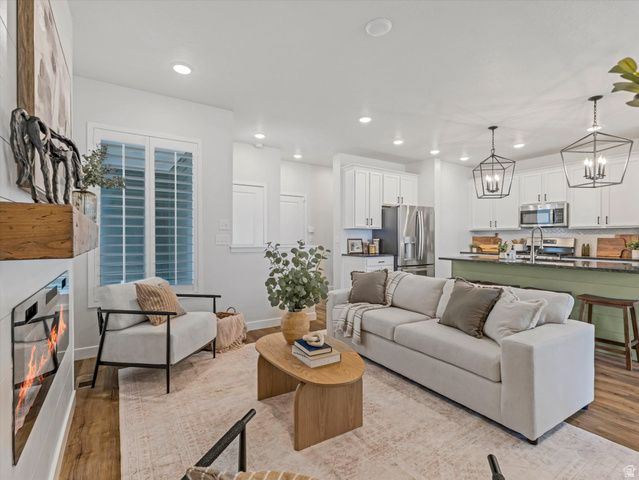 11759 South Watercourse Road South Jordan, UT 84009 - Photo 6 of 33 Open-concept living area featuring wood-finish flooring, a contemporary fireplace with a wood mantle, white cabinetry, a kitchen island with dark countertops, and two geometric pendant lights