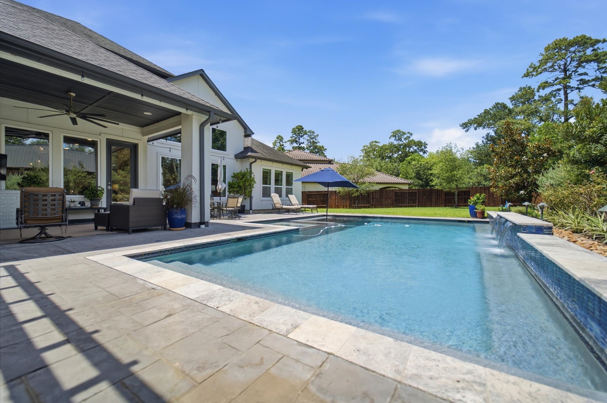 23722 Thortons Park Lane Spring, TX 77389 - Photo 43 of 48 This photo showcases a spacious backyard featuring a modern pool with a waterfall edge, surrounded by a stone patio. The home has a covered outdoor seating area, ideal for relaxation. Lush greenery and trees provide privacy, creating a serene outdoor retreat.