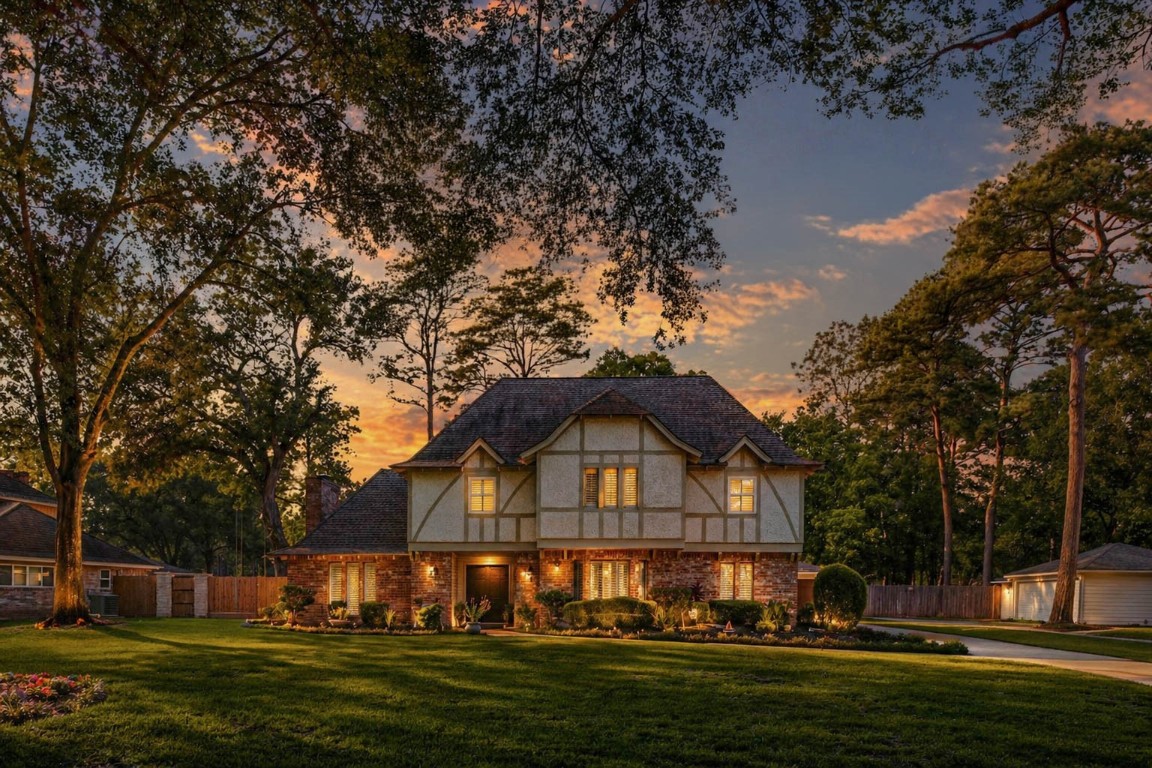 Charming Tudor-style home surrounded by mature trees, with warm, inviting lighting and a well-manicured lawn.