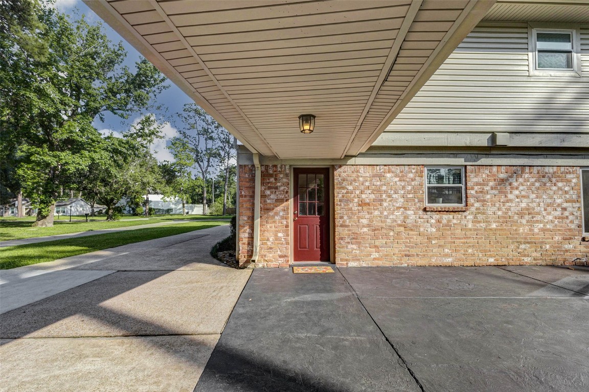 6710 Falling Waters Drive Spring, TX 77379 - Photo 41 of 45 This photo features a home's exterior with a covered entryway and a red door, set against a brick wall. The driveway is spacious, leading up to the entrance, and the surrounding area is landscaped with lush greenery.