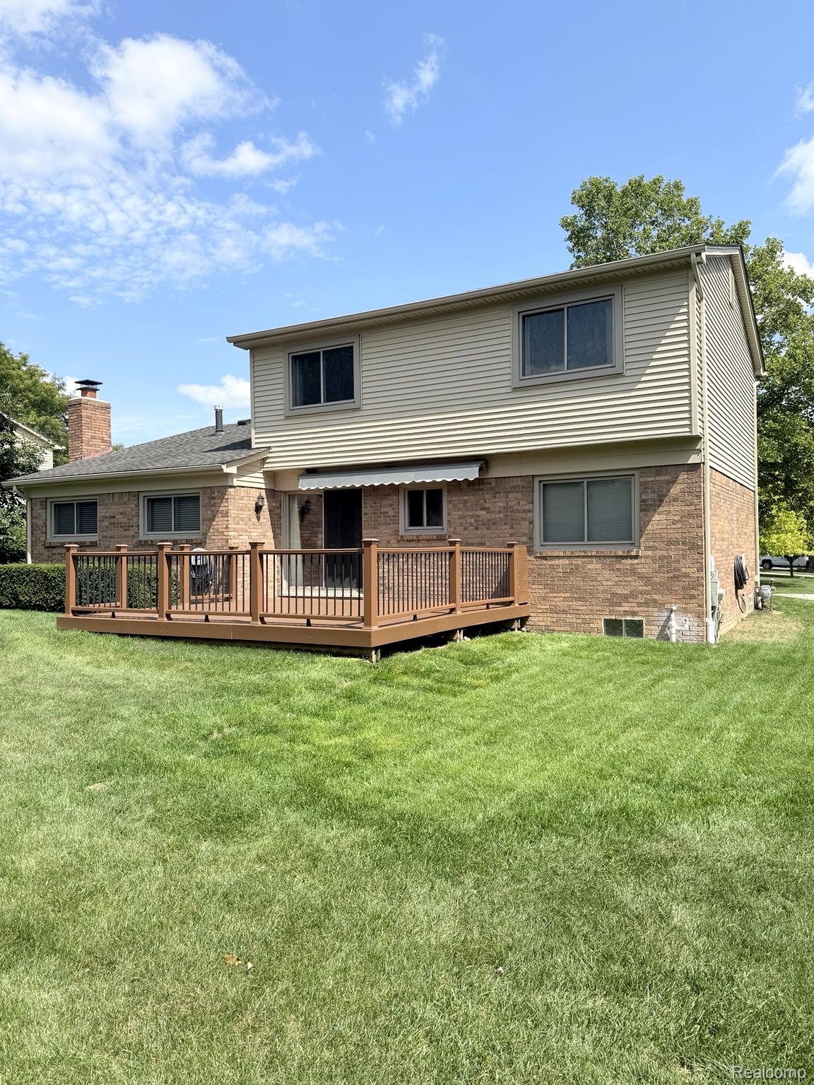 2070 Winchester Road Rochester, MI 48307 - Photo 3 of 34 Expansive backyard featuring a manicured lawn, a two-story brick and siding exterior, a wood-finish deck with dark railings, and an awning over the rear entrance