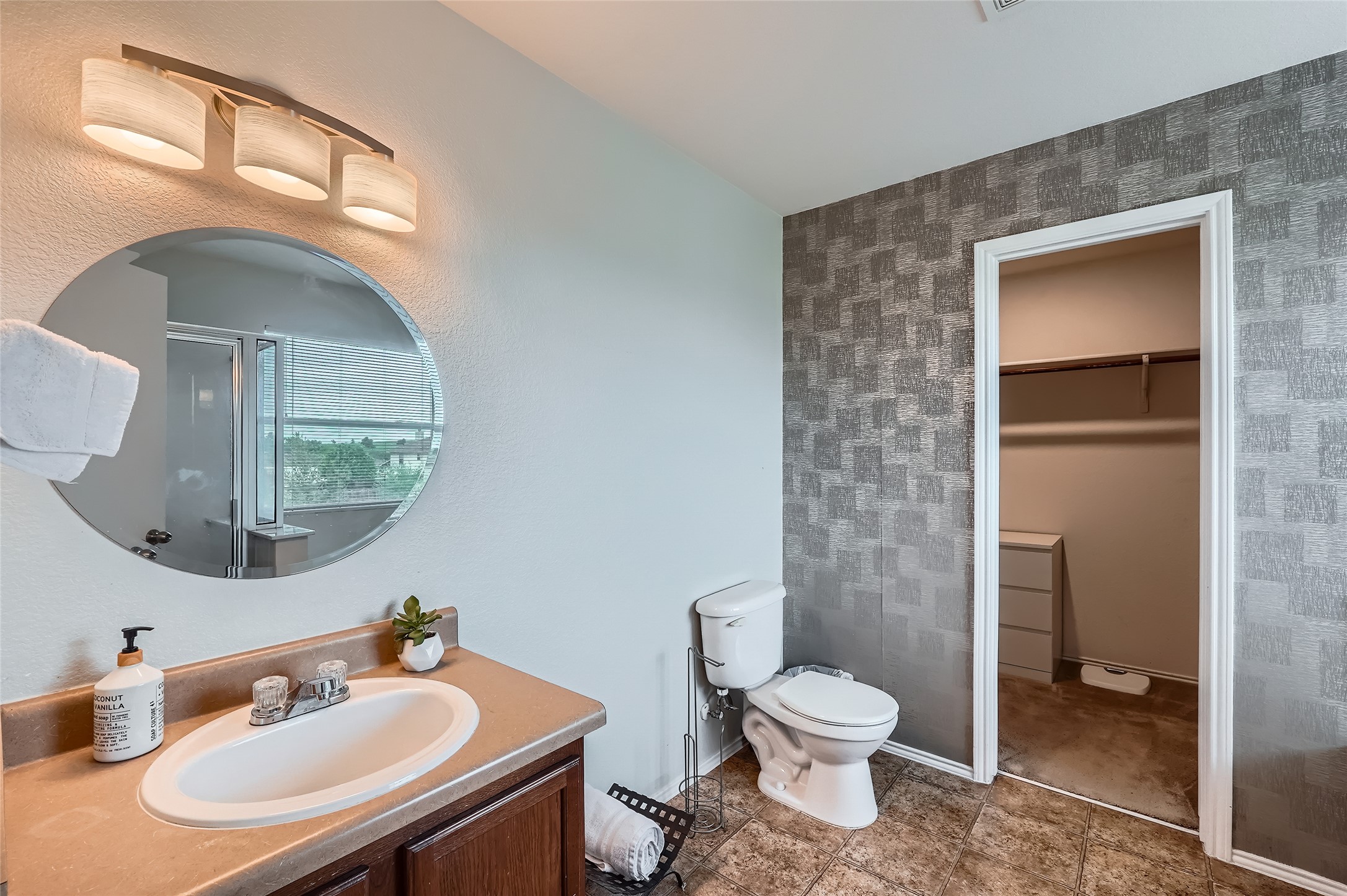 18021 Prairie Verbena Lane Elgin, TX 78621 - Photo 21 of 33 A well-lit bathroom vanity with a mirror reflecting a shower and window, complemented by stylish geometric wallpaper.