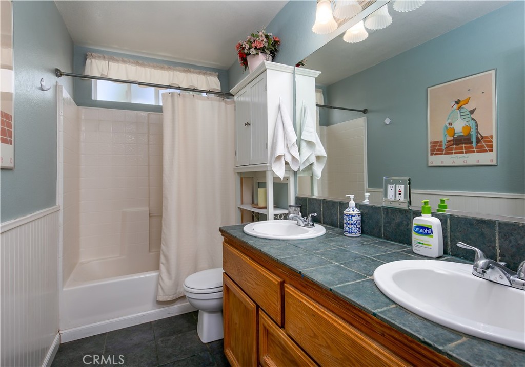 10960 Steinhoff Road Frazier Park, CA 93225 - Photo 35 of 46 Conveniently located upstairs near the secondary bedrooms, this full hallway bathroom offers a long oak vanity with dual sinks, classic fixtures, and custom teal tile countertops. The space includes a clean shower/tub combination with a white curtain, a standard commode, and a soft blue-grey accent wall.