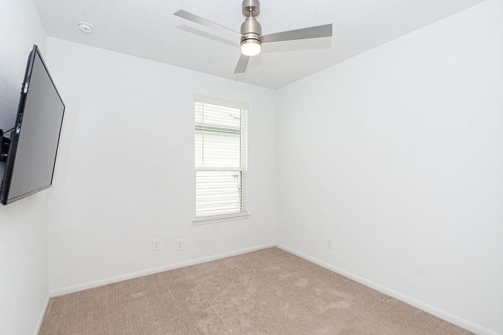 23502 Glenbuck St Spring Spring, TX 77373 - Photo 38 of 50 This room features a clean, minimalist design with white walls, a ceiling fan, carpeted floor, and a wall-mounted TV. The window allows natural light to brighten the space, making it suitable for a cozy bedroom or home office.