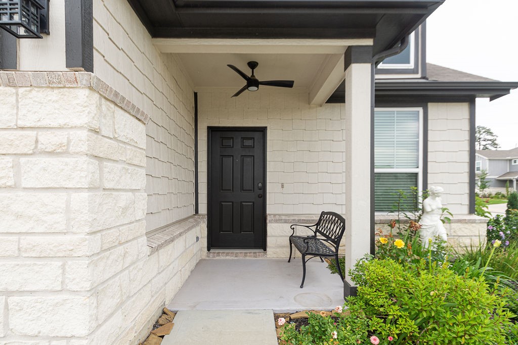 23502 Glenbuck St Spring Spring, TX 77373 - Photo 5 of 50 This photo showcases a charming front porch with a black door, a ceiling fan, and a decorative bench. The area is framed by light-colored brick and neatly landscaped with flowers and a small statue, offering a welcoming entrance to the home.