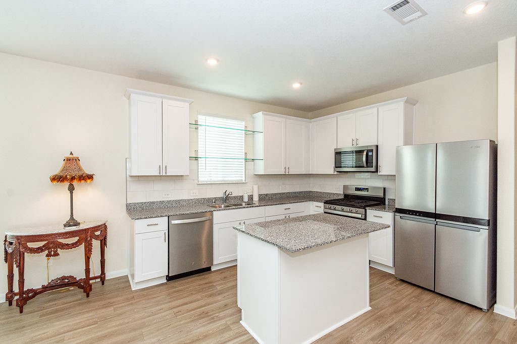 23502 Glenbuck St Spring Spring, TX 77373 - Photo 8 of 50 This modern kitchen features sleek white cabinets, granite countertops, and stainless steel appliances, including a double-door refrigerator and built-in microwave. An island provides additional workspace, and the space is enhanced by ample lighting and a stylish decorative table with a lamp.