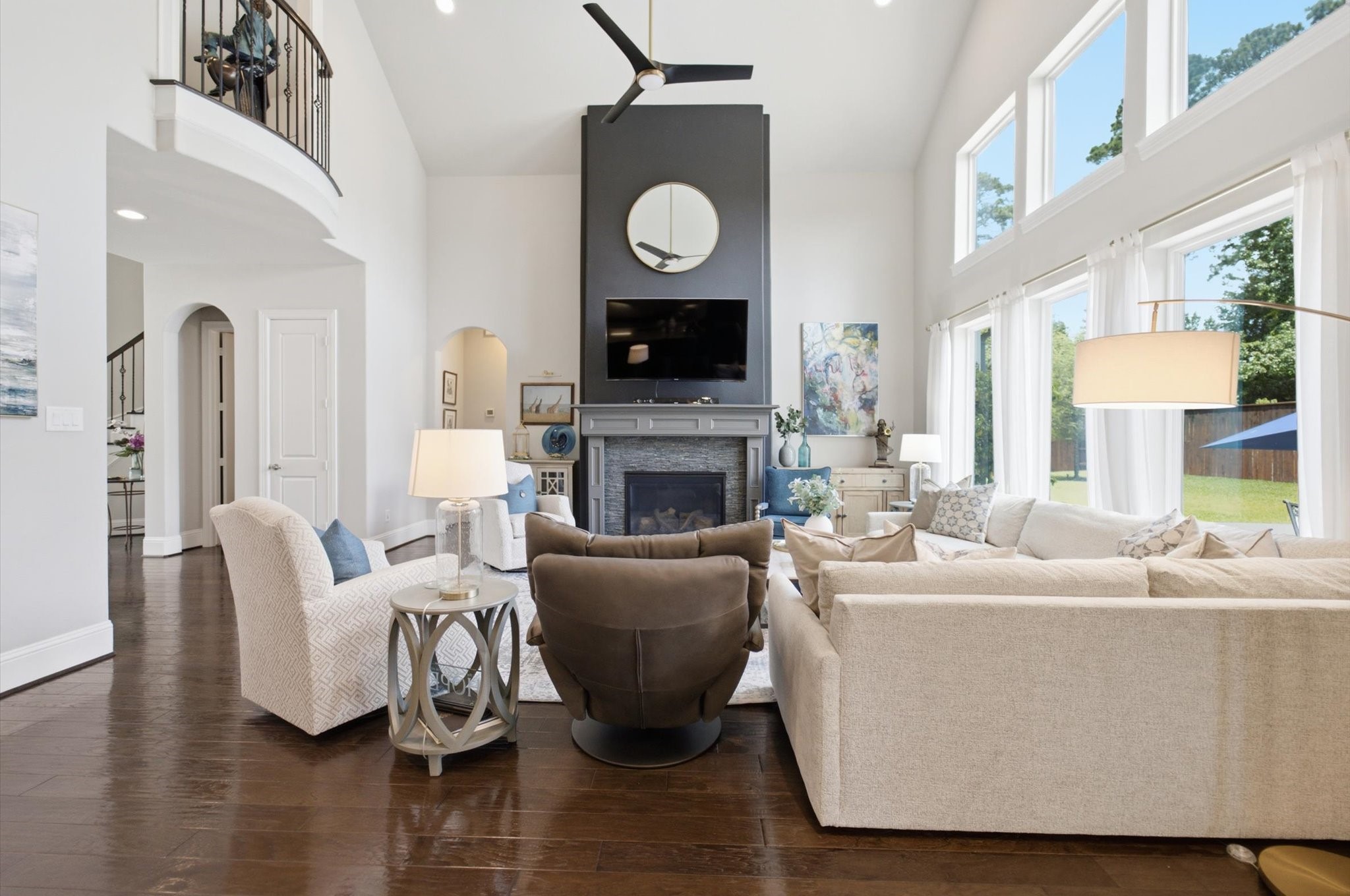 23722 Thortons Park Lane Spring, TX 77389 - Photo 28 of 48 This photo showcases a spacious, elegant living room with high ceilings and abundant natural light from large windows. It features a cozy seating arrangement, a modern fireplace with a mounted TV above, and stylish decor. The room has a contemporary feel with light-colored walls, dark hardwood floors, and a view of a lush backyard.