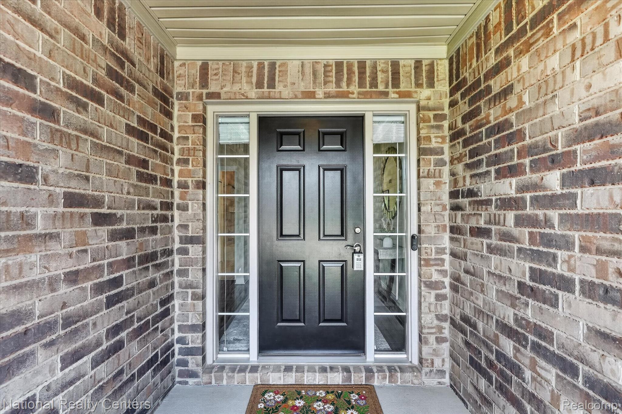 740 North Mill Street South Lyon, MI 48178 - Photo 9 of 56 Panel-style entry door with sidelights, complemented by a brick façade and a concrete porch