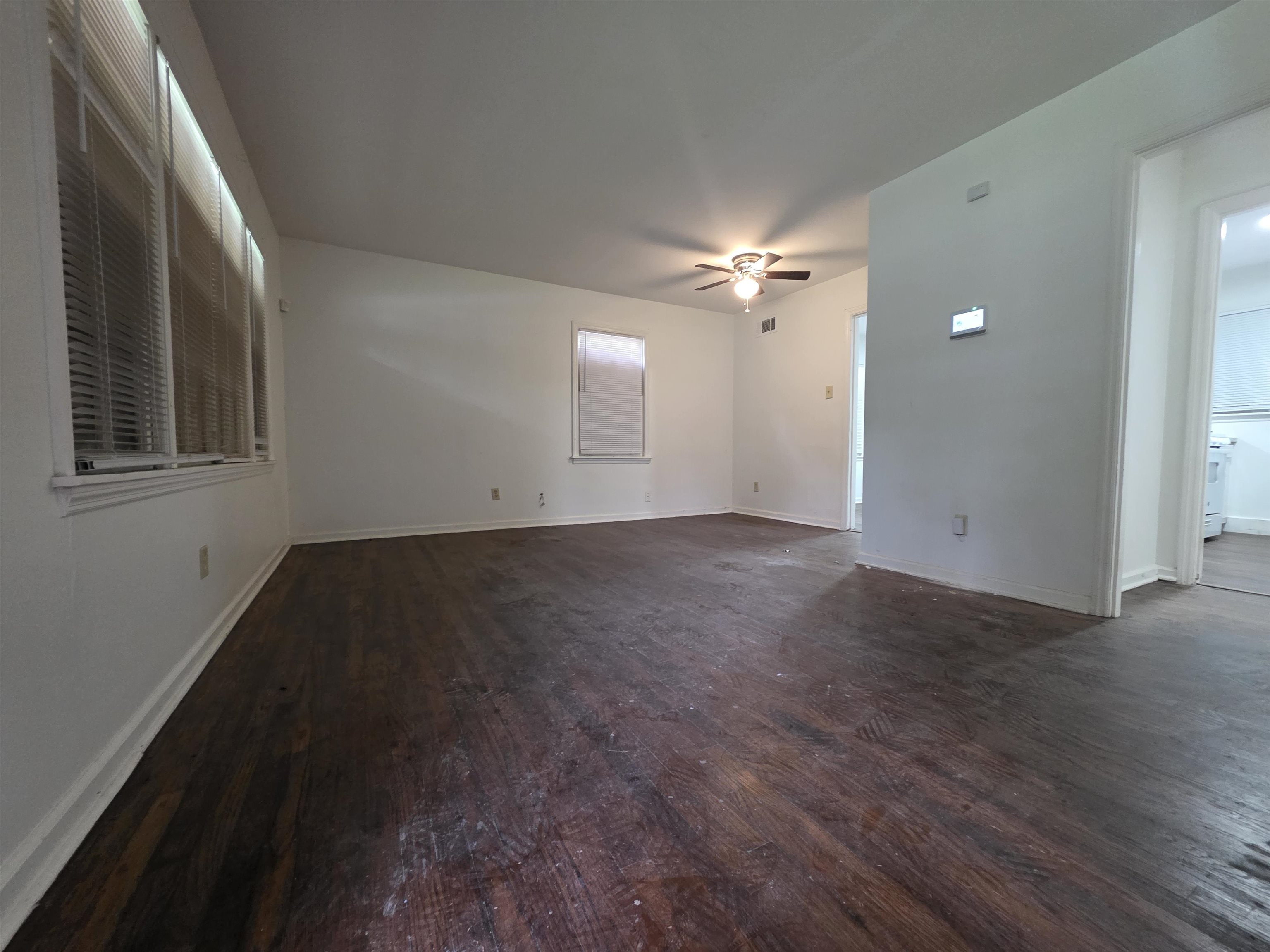 Spacious interior featuring wood-finish flooring, white walls, multiple windows with horizontal blinds, and a ceiling fan with integrated lighting