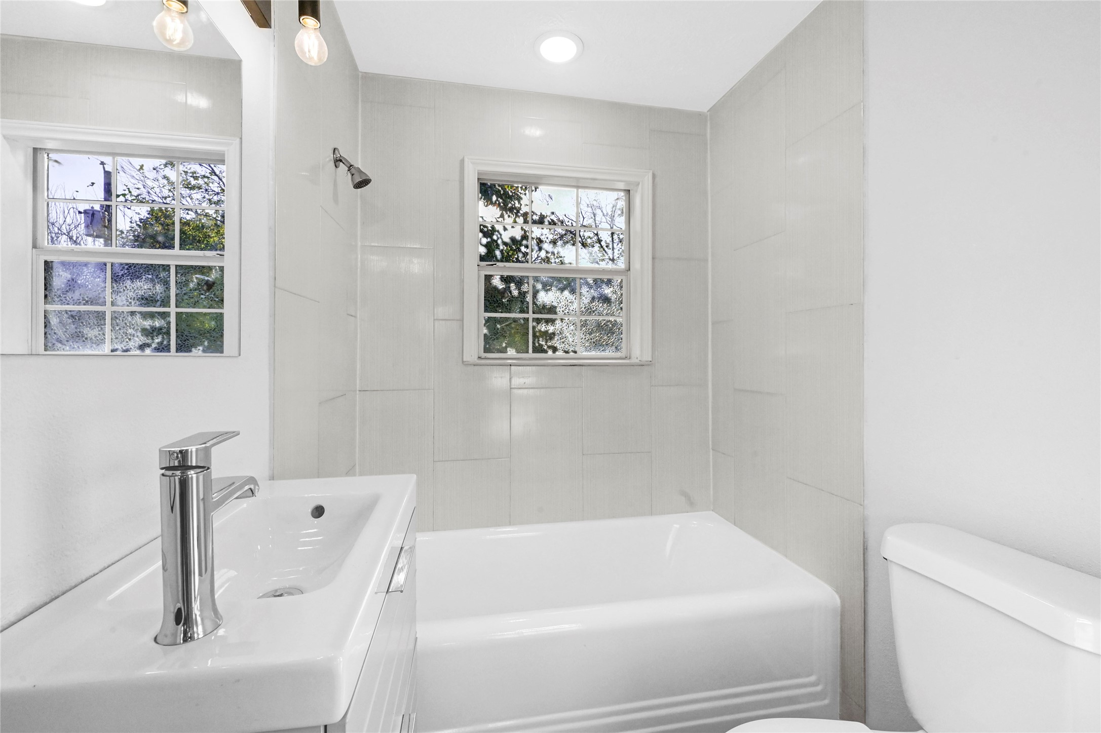 1633 Fourcade Street, Unit C Houston, TX 77023 - Photo 6 of 7 Bright, modern bathroom featuring a sleek white sink and vanity, bathtub, and toilet. Natural light streams through two windows, enhancing the clean, minimalist design.