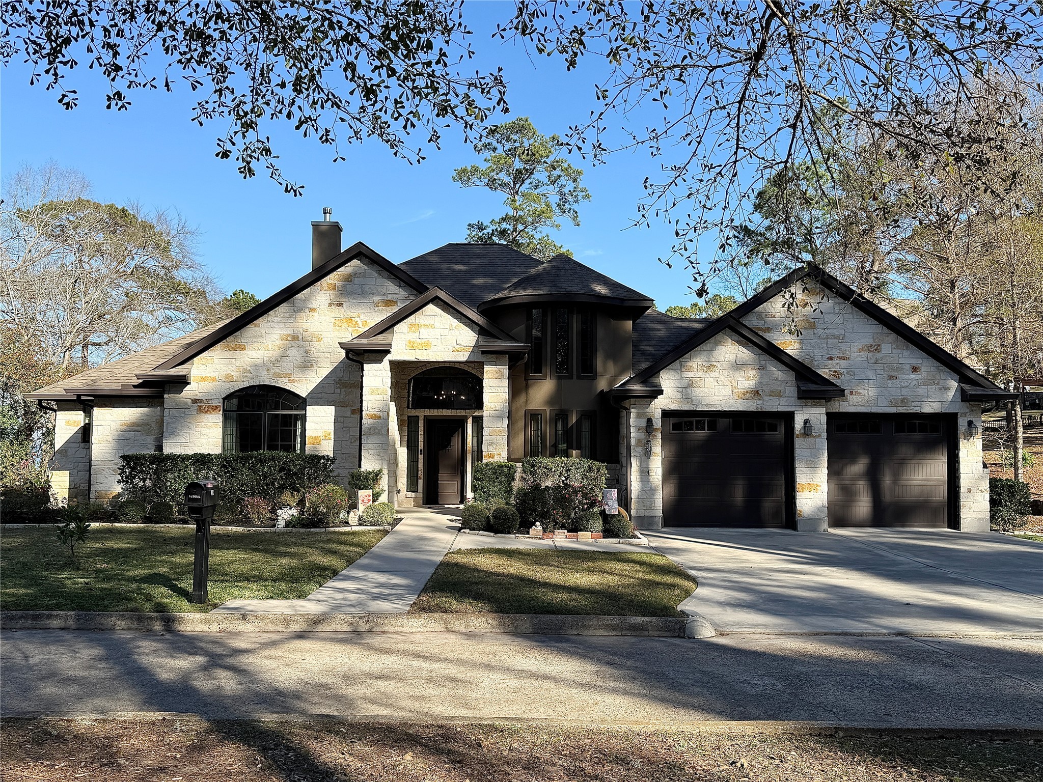 This photo showcases a charming, single-story stone house with a dark shingled roof, featuring a two-car garage and a welcoming front entrance. The beautifully landscaped front yard adds to its curb appeal, set against a backdrop of mature trees.