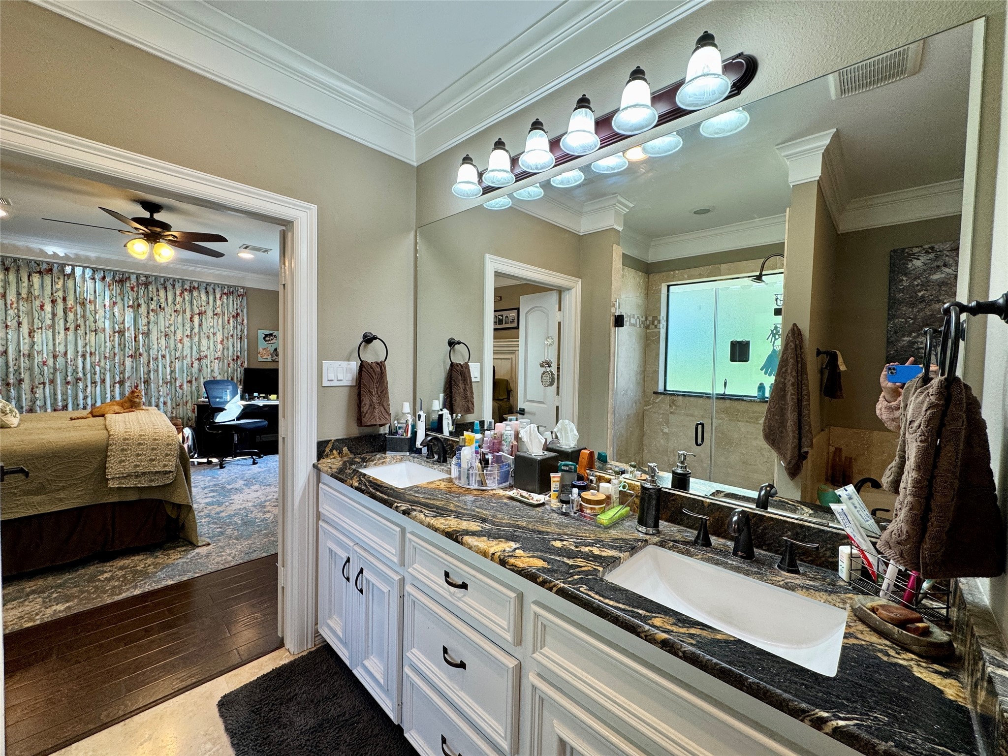 200 Harbor Run Drive Coldspring, TX 77331 - Photo 29 of 50 This photo showcases a spacious primary bathroom with a dual-sink vanity featuring elegant granite countertops. The room has ample lighting, a large mirror, and a glass-enclosed shower. It opens into a large bedroom with a ceiling fan, engineered wood floors, and floral curtains.